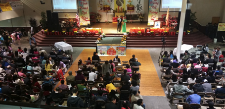 A crowded indoor event with a stage, audience seated in chairs, and Christmas decorations.