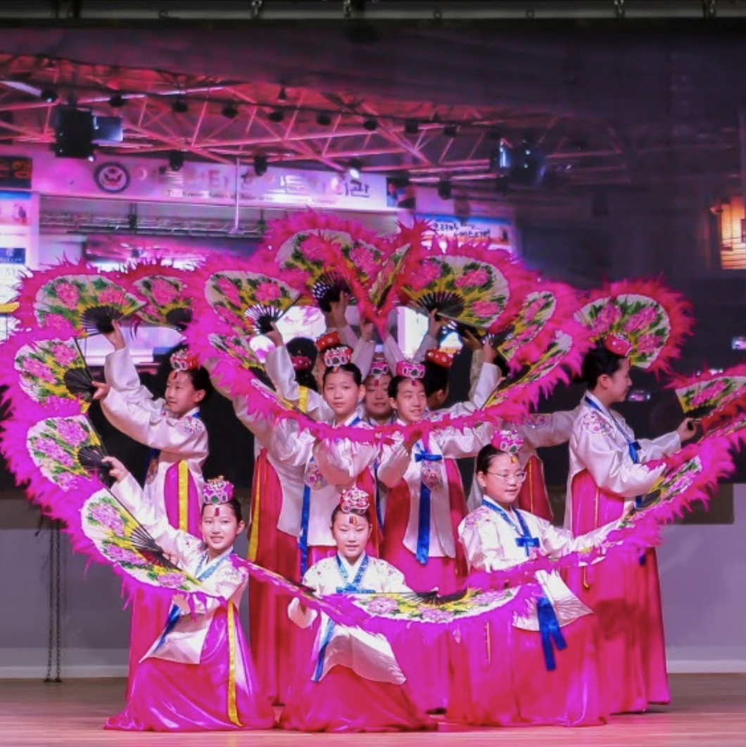 Group of young girls in traditional Korean hanbok performing a dance with large pink feathered fans on stage, colorful costumes, and a background with stage lighting and banners.