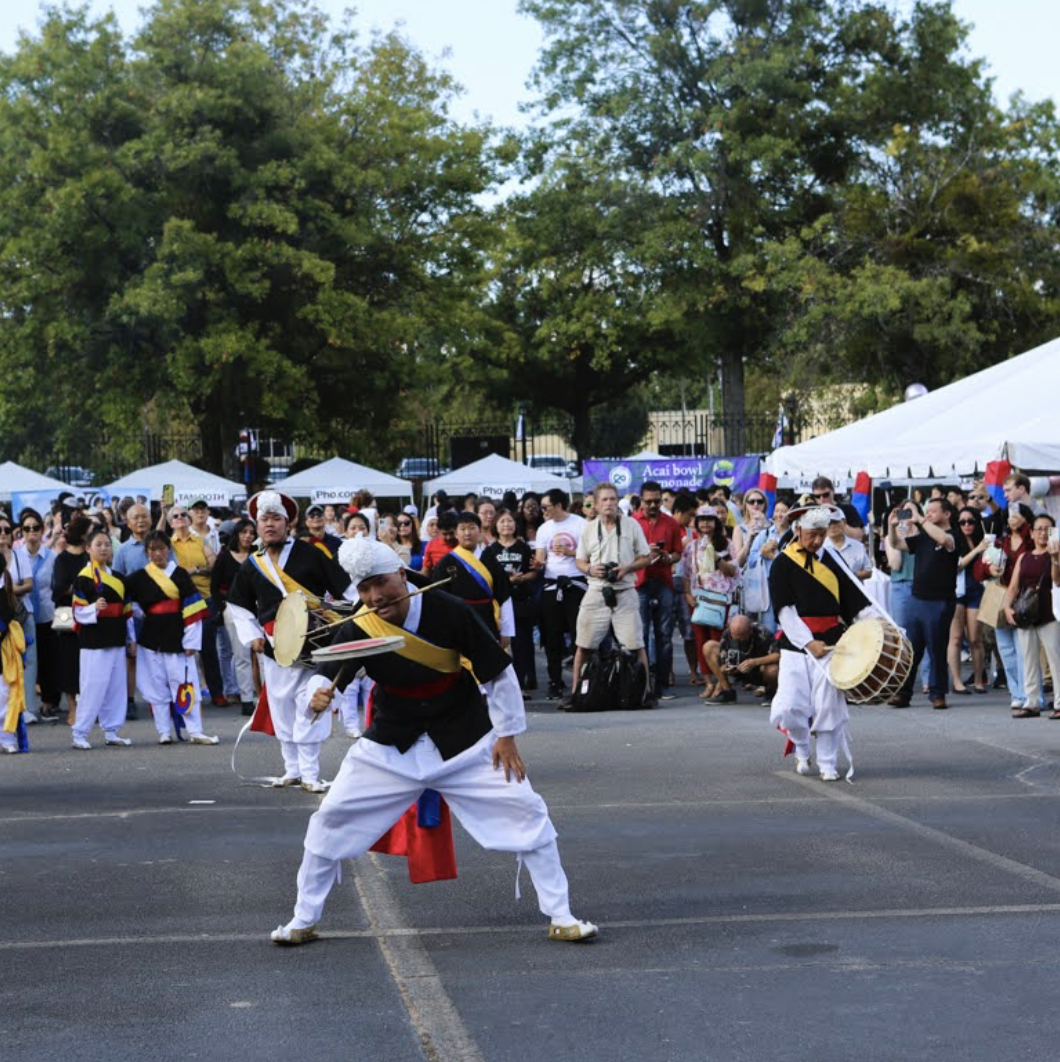 Traditional Korean performers in colorful costumes performing a dance with props in front of an audience at an outdoor festival. There are tents and trees in the background.