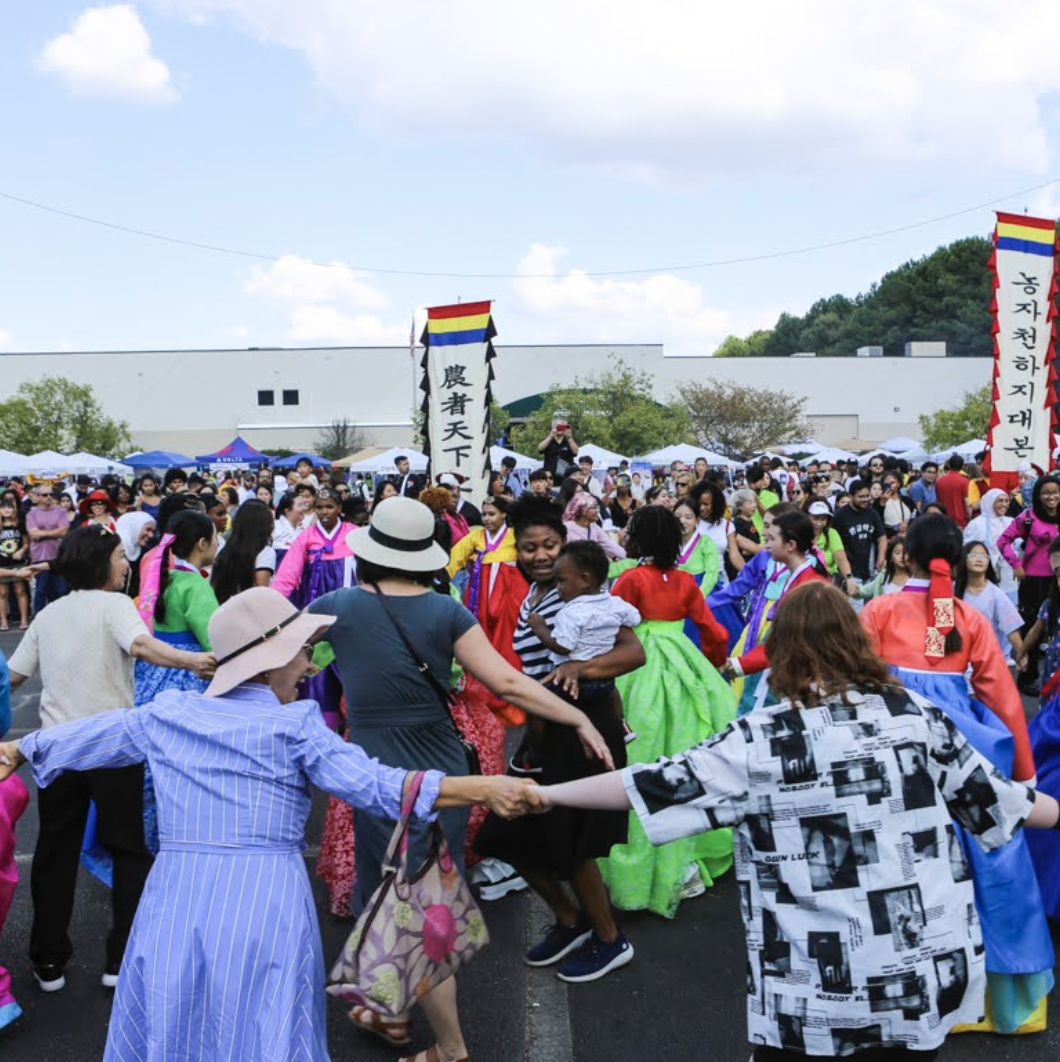 People dancing in traditional Korean hanboks during an outdoor festival with tents and banners.