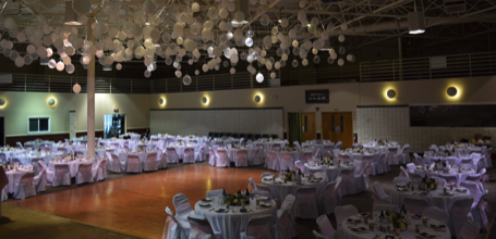 Large banquet hall decorated for an event with white draped ceilings and round tables covered with white tablecloths and chairs, set with tableware and centerpieces.