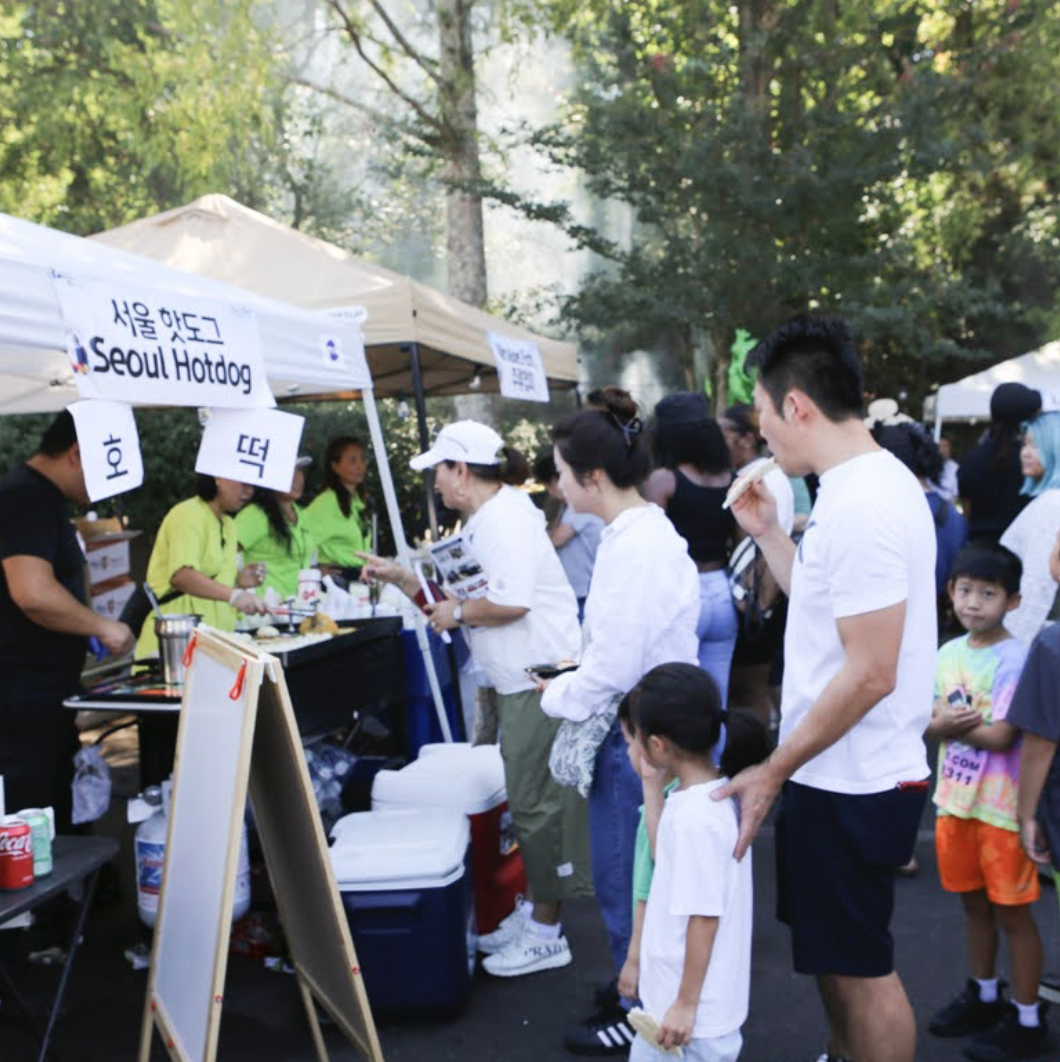 People line up at a Seoul Hotdog stand at an outdoor park event with trees in the background.