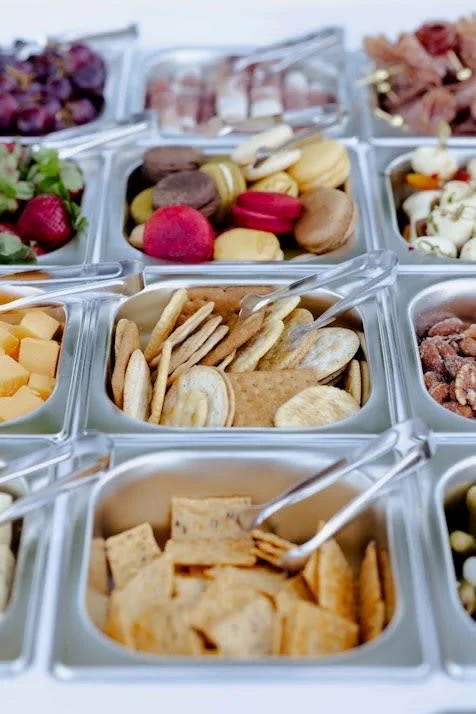 Assorted items in stainless steel containers, including cheese cubes, crackers, cookies, grapes, strawberries, and various finger foods.