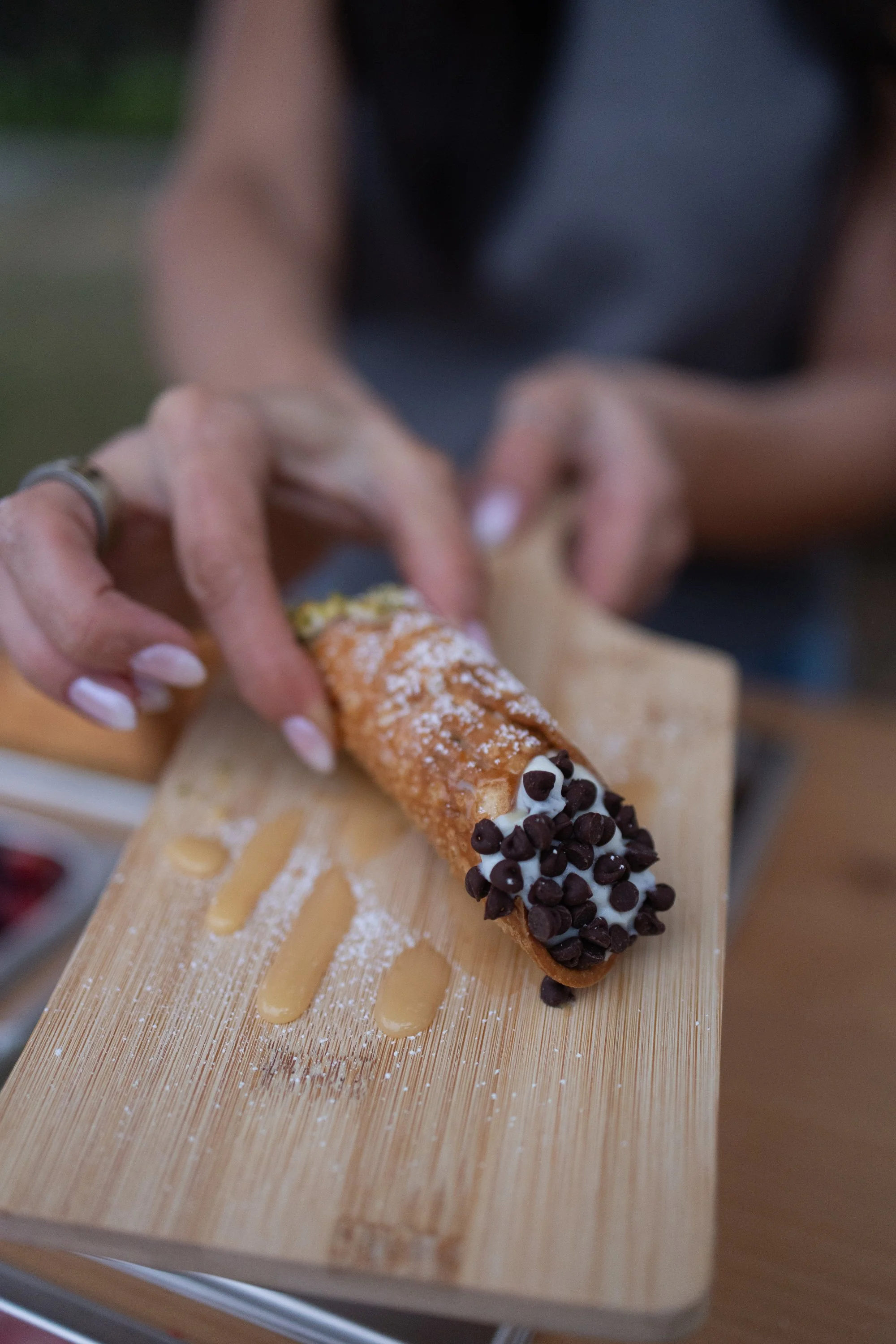 Person holding a cannoli with chocolate chips and whipped cream on a wooden serving board