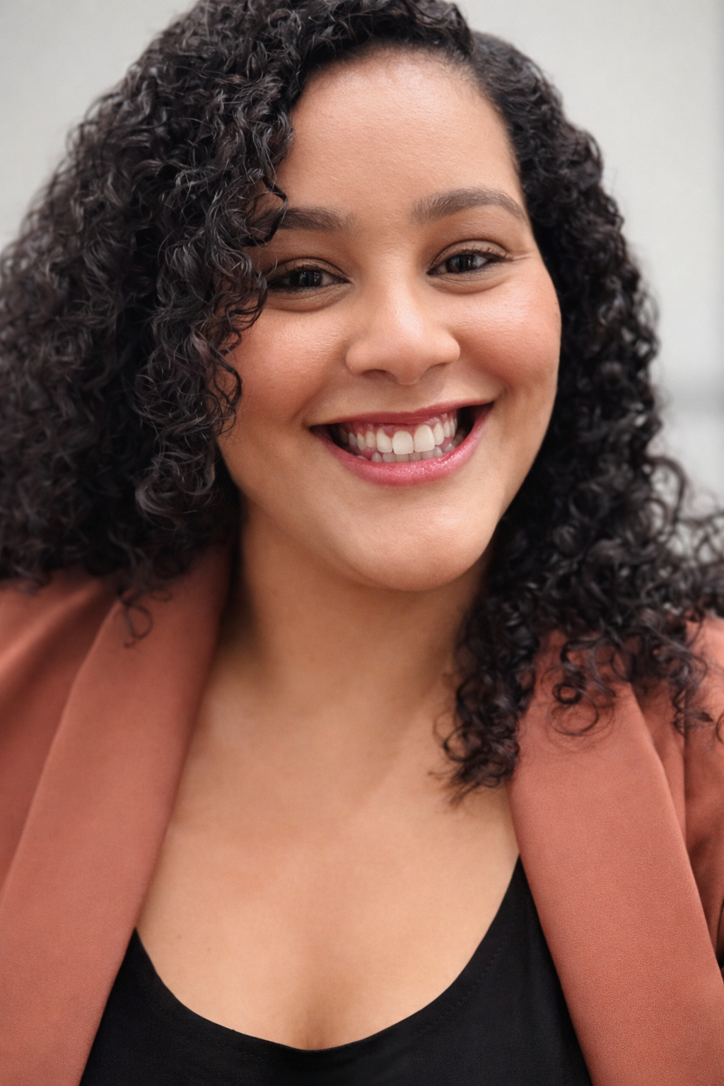 Close-up of a smiling woman with curly dark hair, wearing a brown jacket and black top.