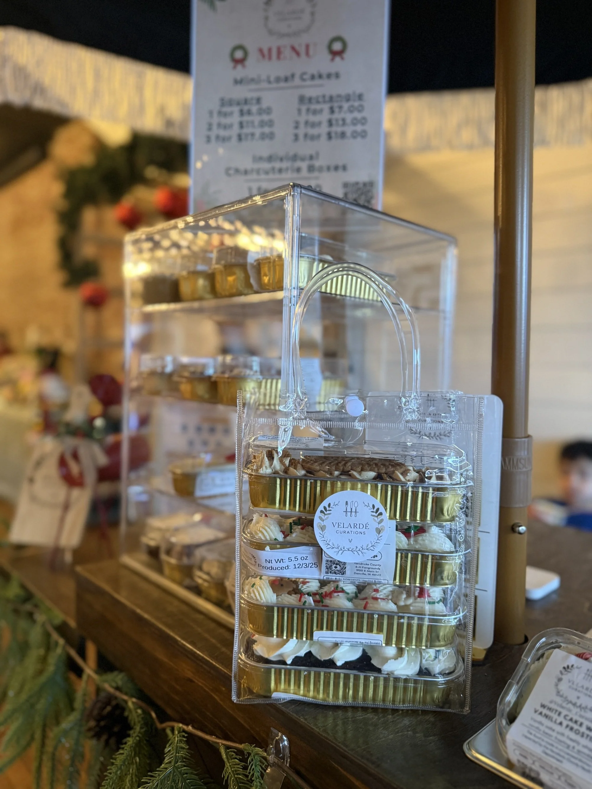 Clear acrylic display case with mini cheesecake slices on a wooden table at a holiday-themed bakery or bakery event. A holiday menu is visible in the background.