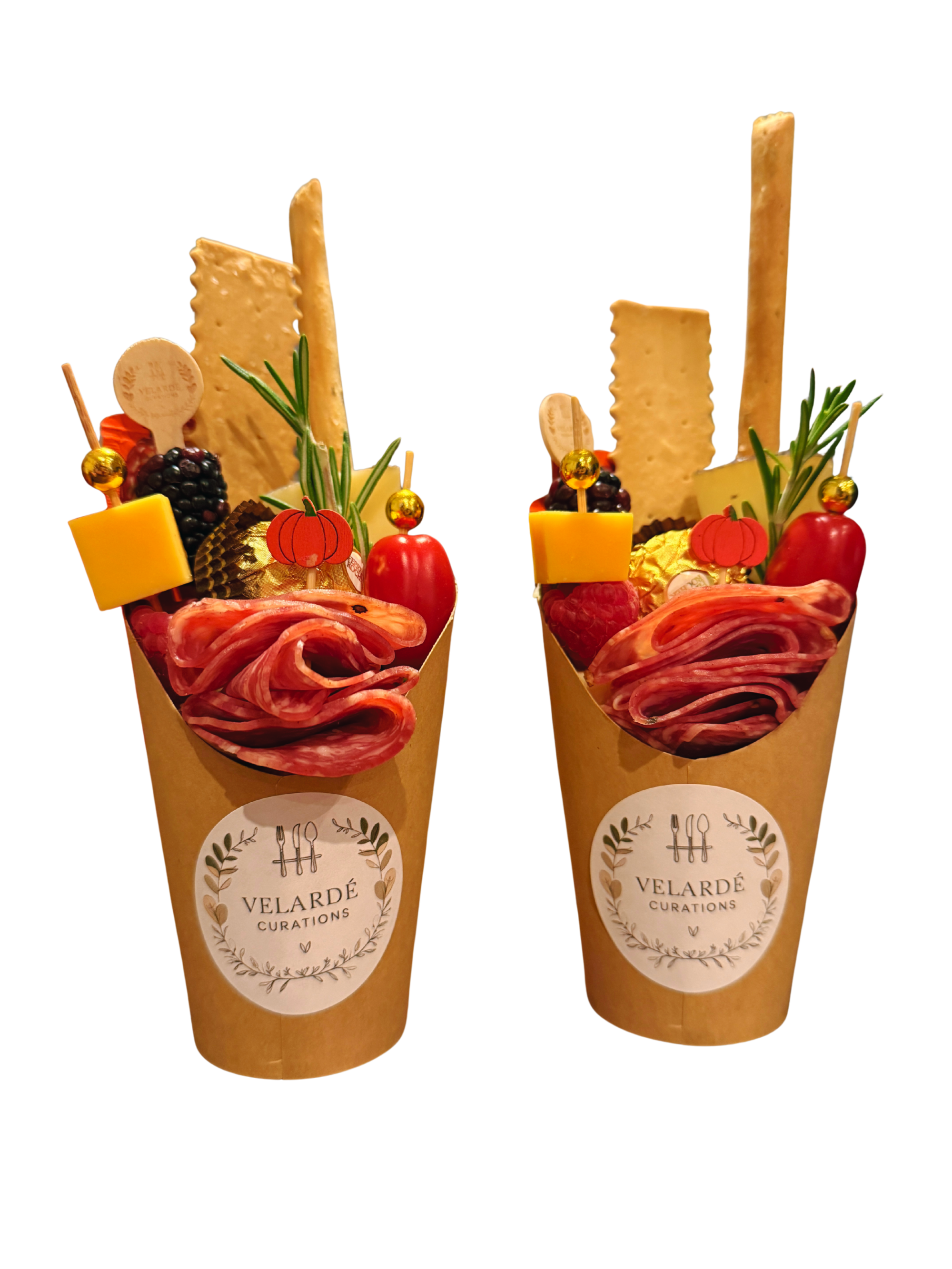 Two charcuterie cups with cured meats, cheese, crackers, cherry tomatoes, berries, herbs, and decorative elements.