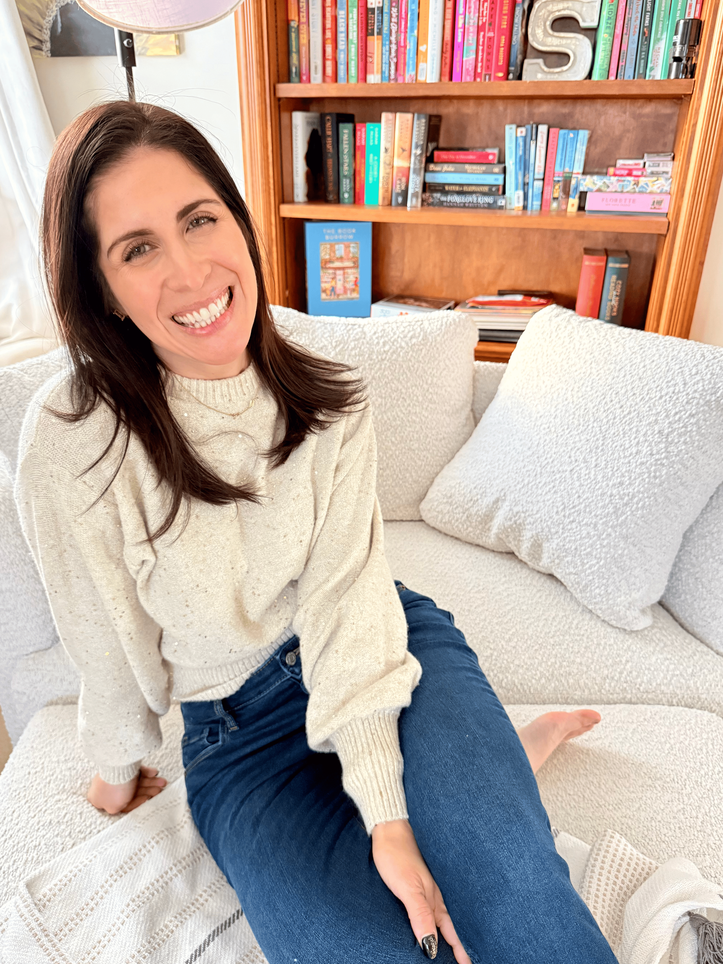 A woman with dark brown hair smiling, sitting on a beige couch in a room with bookshelf behind her, wearing a cream sweater and blue jeans.