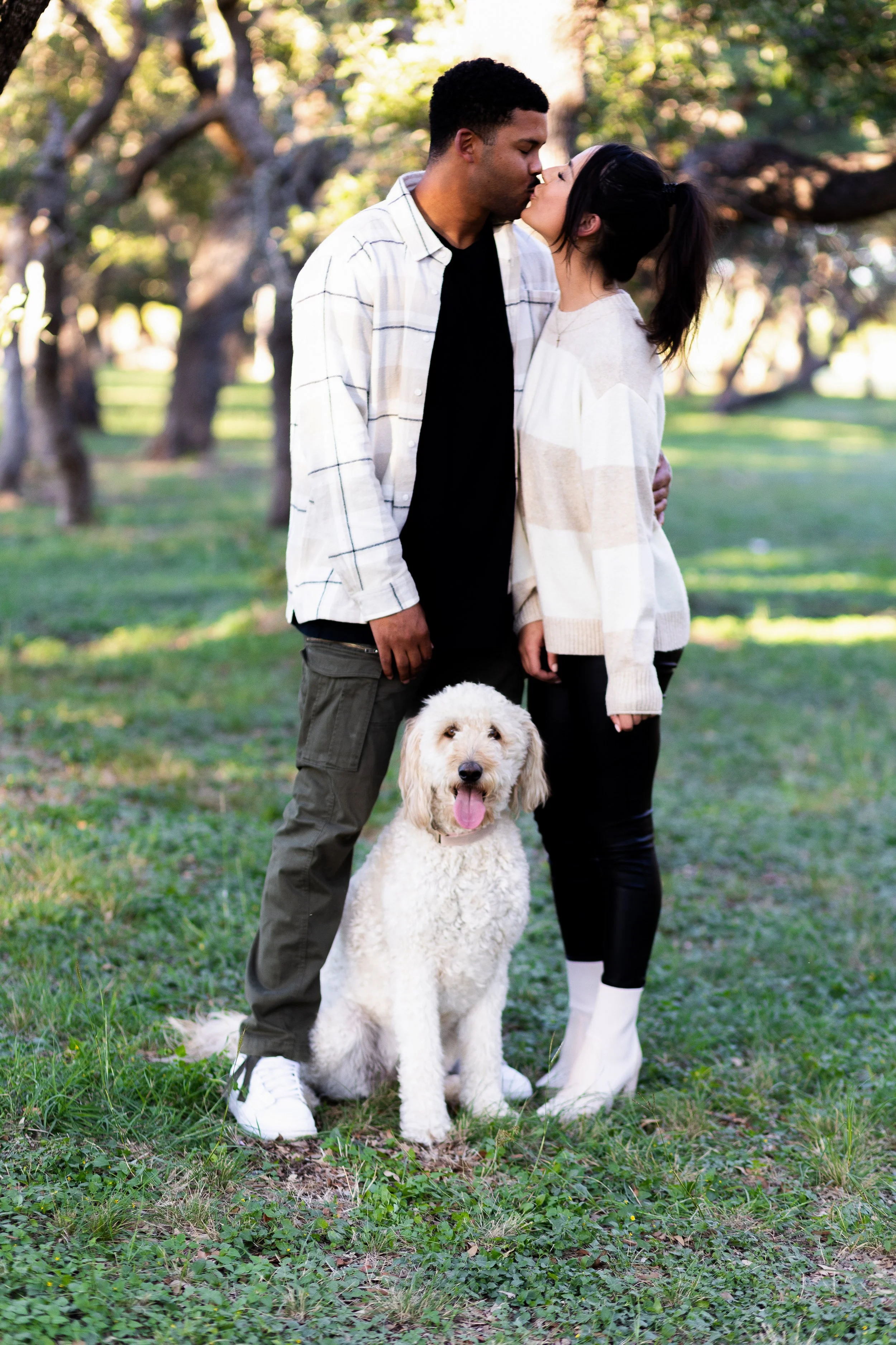 A couple kisses outdoors in a park, with their dog sitting in front of them.