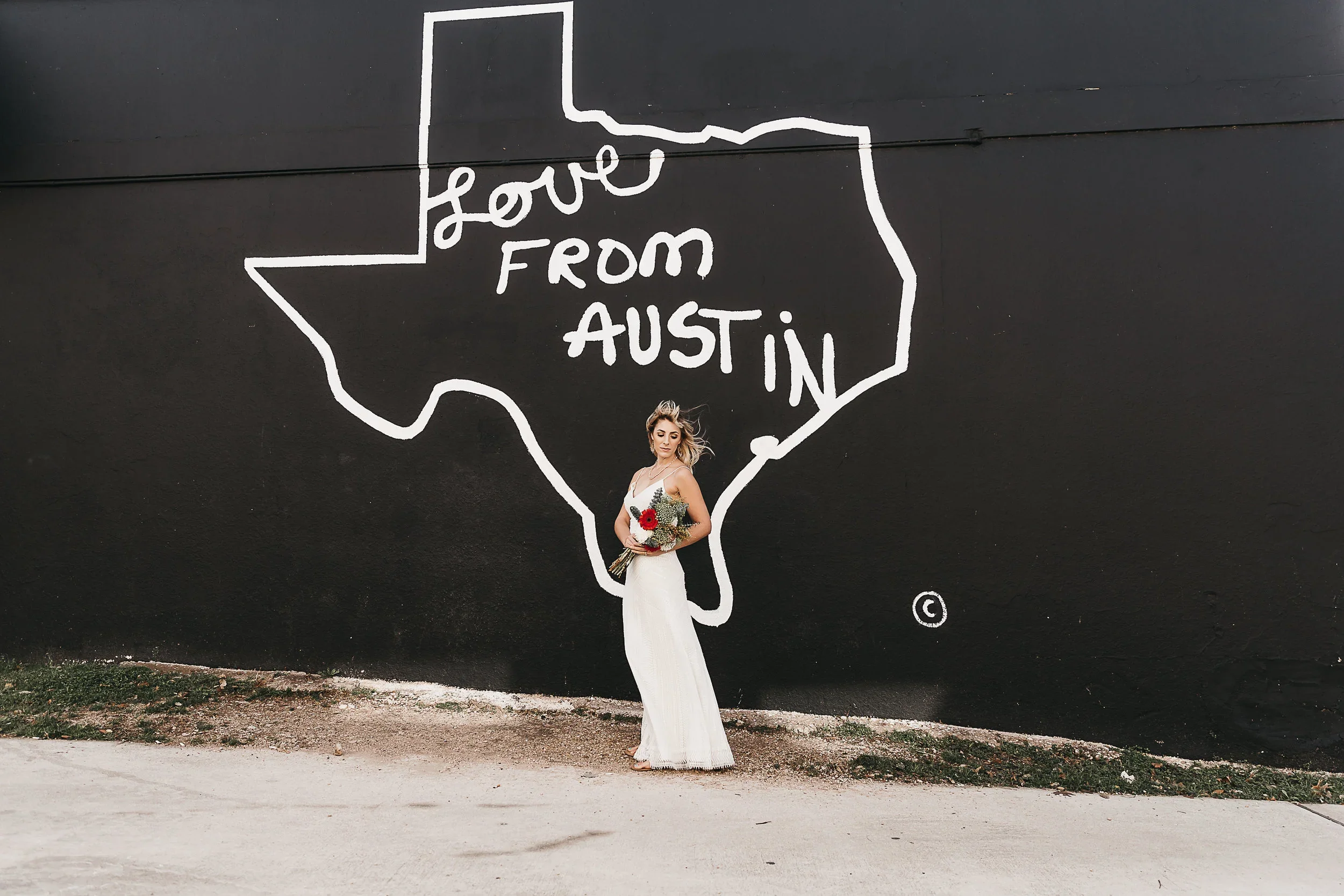 Woman in a long white dress holding a bouquet of flowers standing in front of a black wall with a white outline map of Texas and the words "Love from Austin."