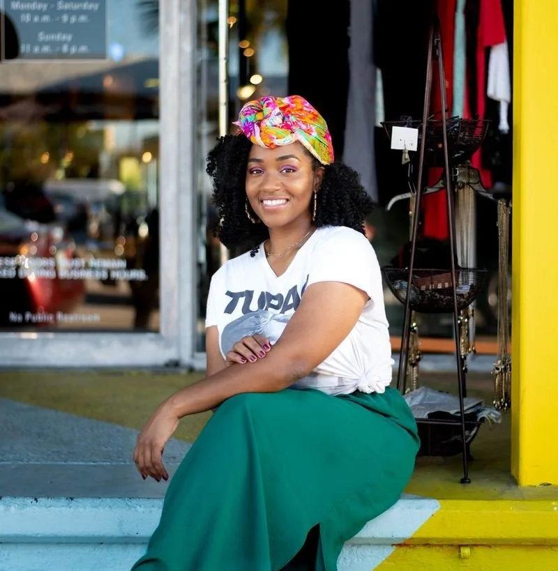 A woman sitting outside modeling, wearing a colorful headscarf, white T-shirt, and green skirt, smiling at the camera, with a store window and clothes rack in the background.