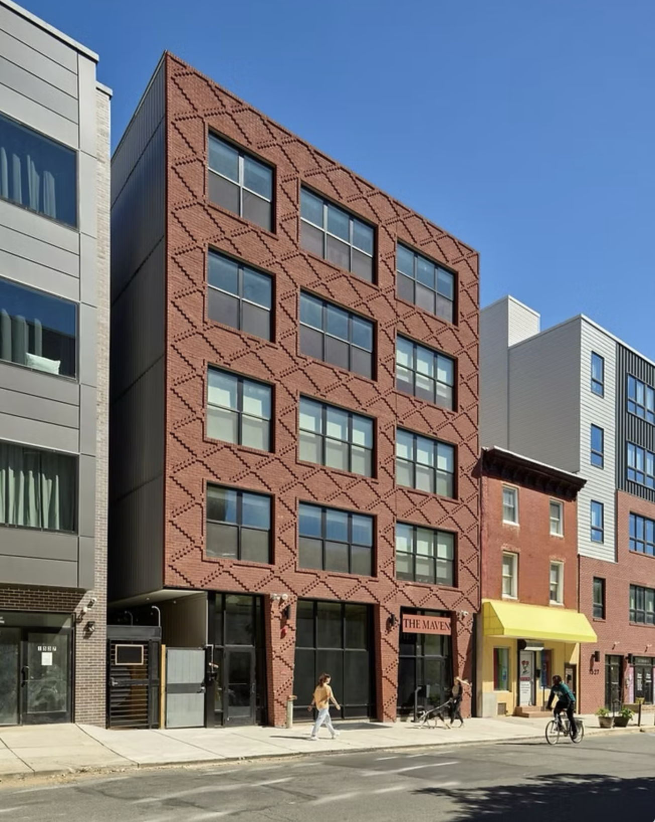 Multi-story modern building with a distinctive red brick facade, large windows, and a sign that says 'The Maven' at street level, with people walking and biking on the sidewalk in front under a clear blue sky.