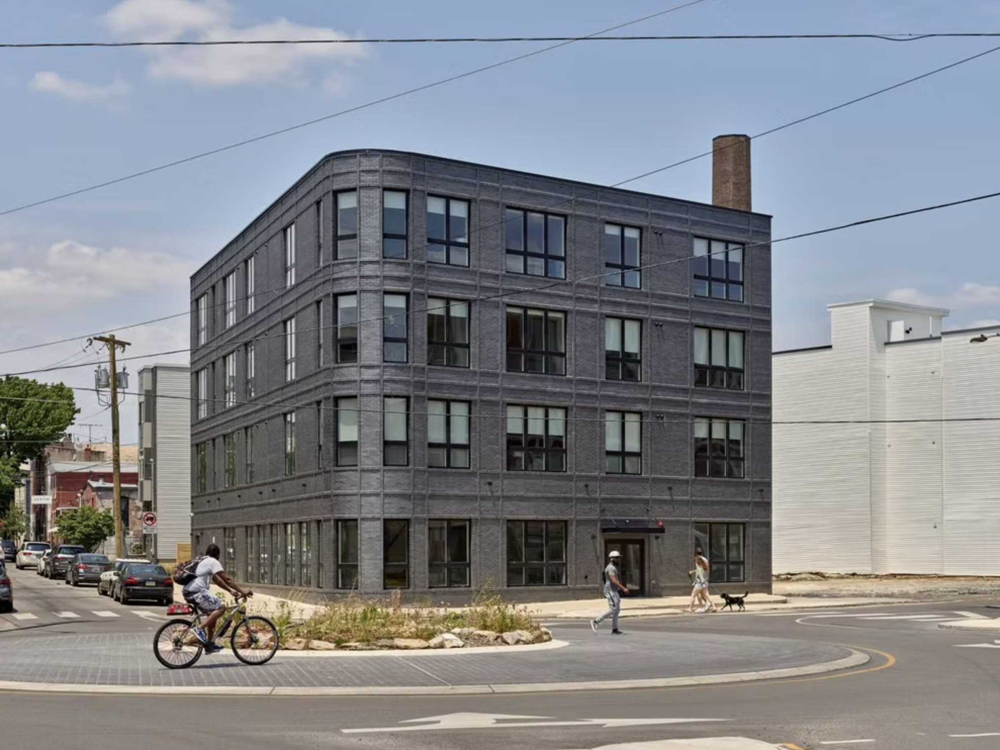 A modern four-story building with dark brick exterior and large windows situated at a street corner, with pedestrians and a cyclist in front.