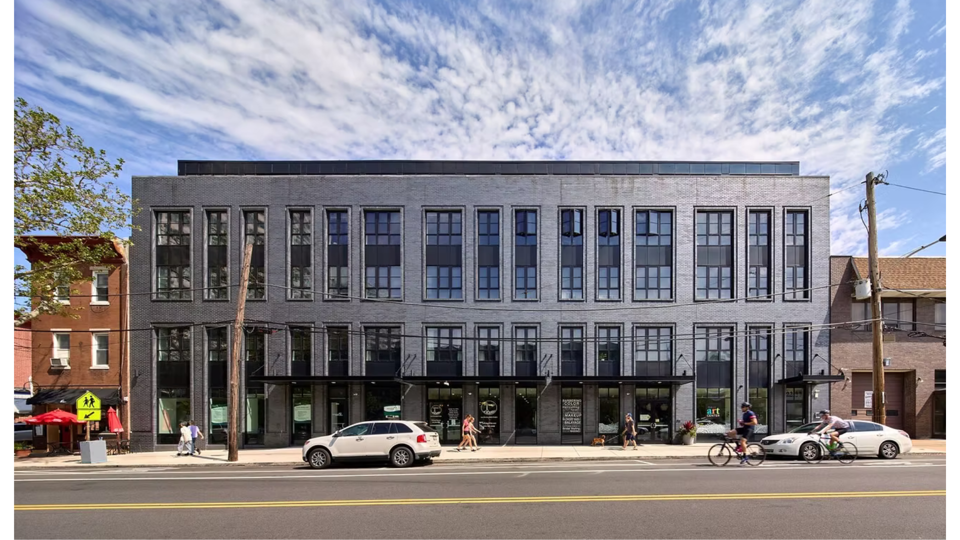 A modern multi-story gray brick building with large grid-style windows on a city street. Pedestrians and cyclists are in front, with parked cars on the street and utility poles visible. The sky is partly cloudy.