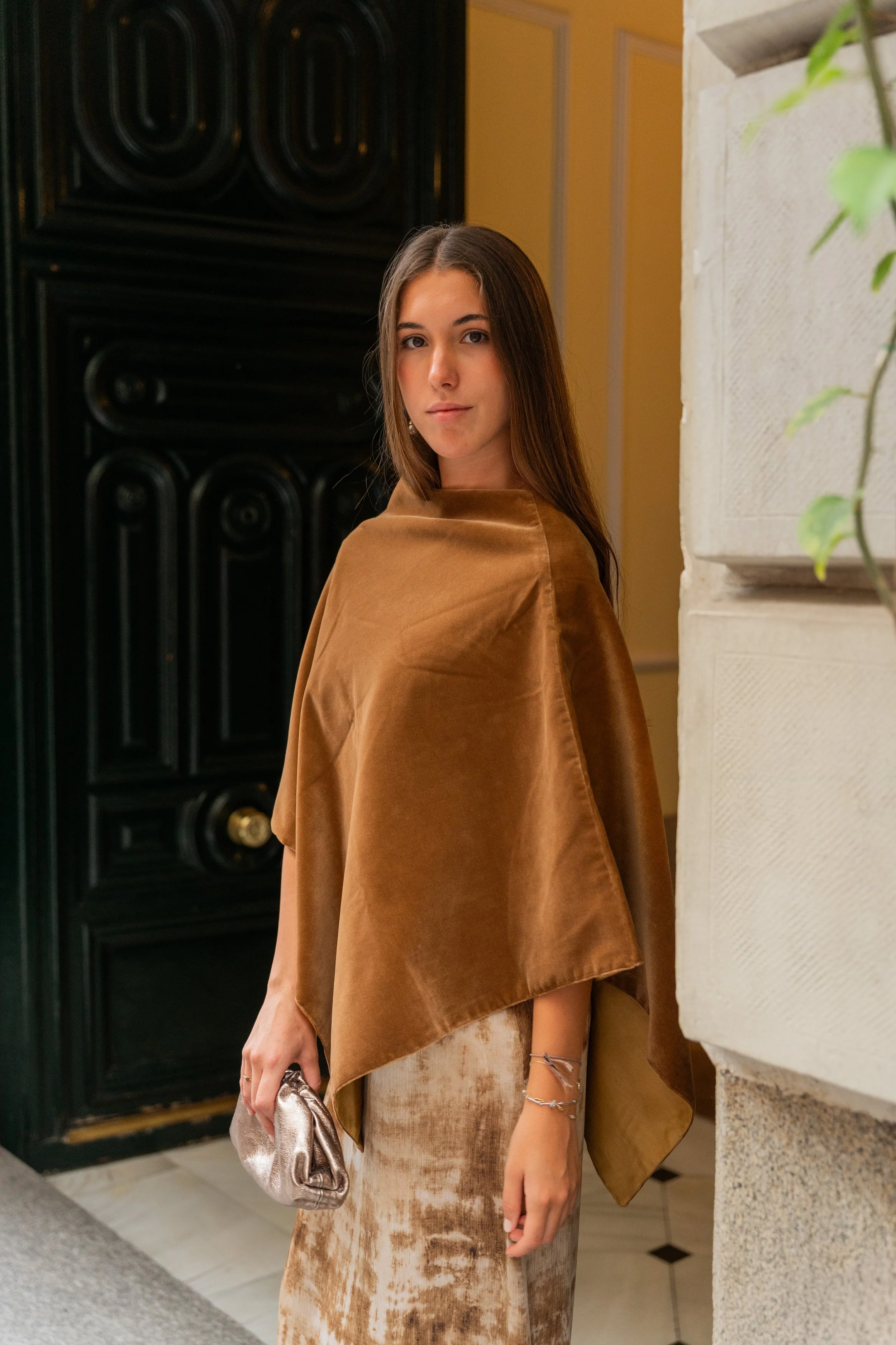 A young woman standing indoors near a stone wall, wearing a brown velvet poncho, a matching gown, holding a shiny silver clutch, and wearing jewelry, with a black decorative door and a yellow wall in the background.