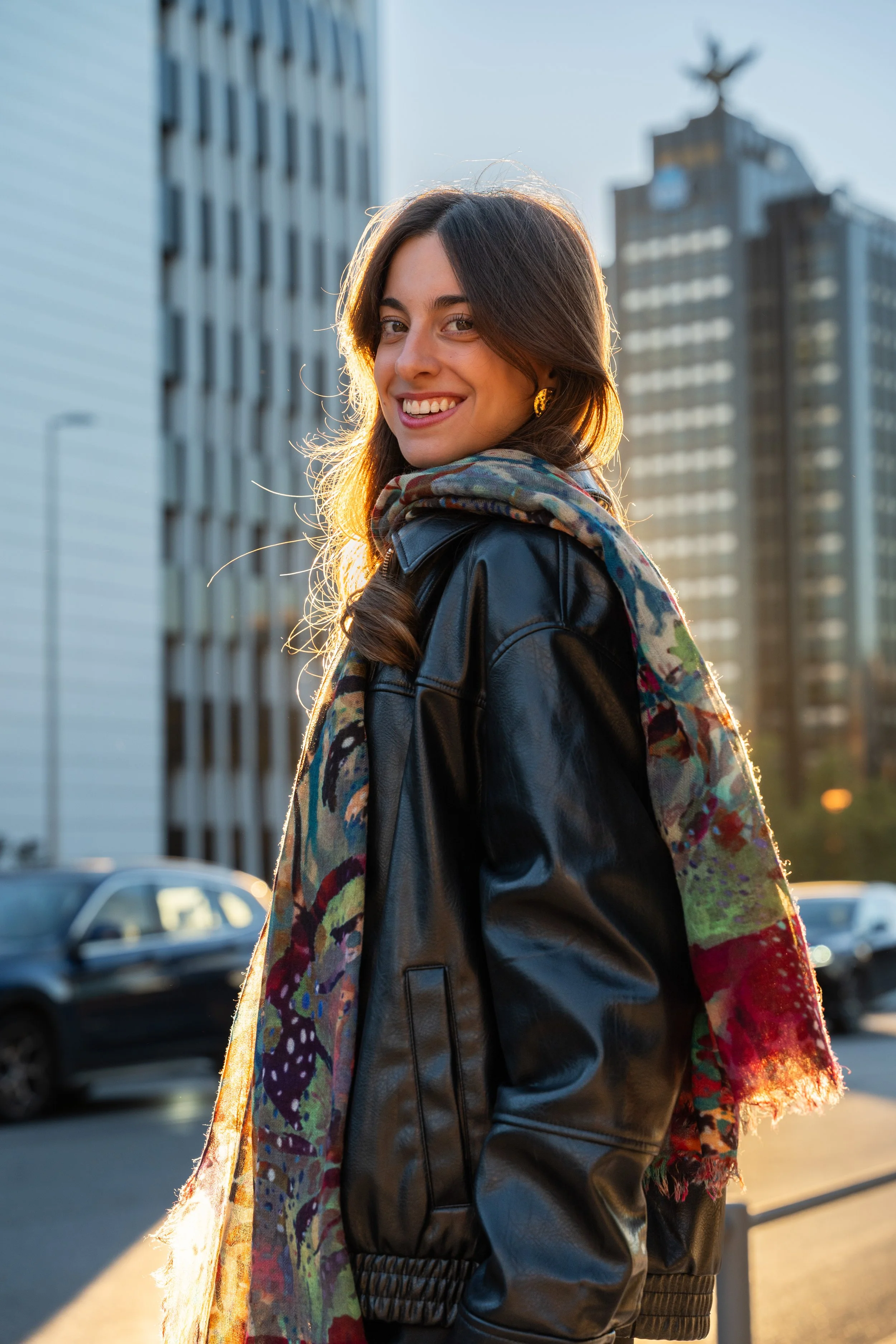 A young woman with long brown hair, wearing a black leather jacket and a colorful scarf, smiling in an urban setting during sunset with tall buildings and parked cars in the background.