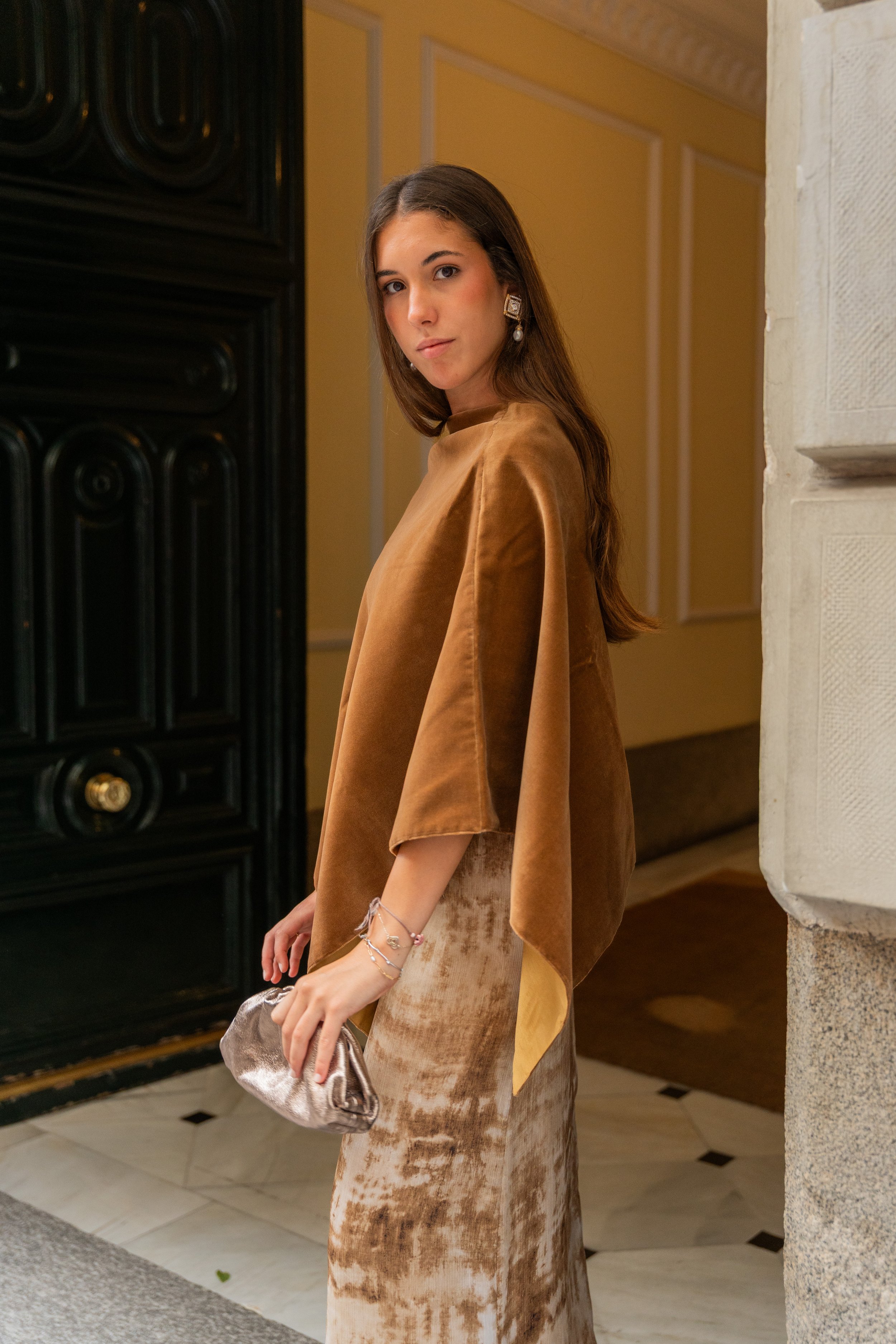 A young woman with long brown hair in an elegant indoor setting. She is wearing a shiny brown top, a patterned beige and brown skirt, gold earrings, and a bracelet. She holds a metallic silver clutch and looks at the camera with a neutral expression.
