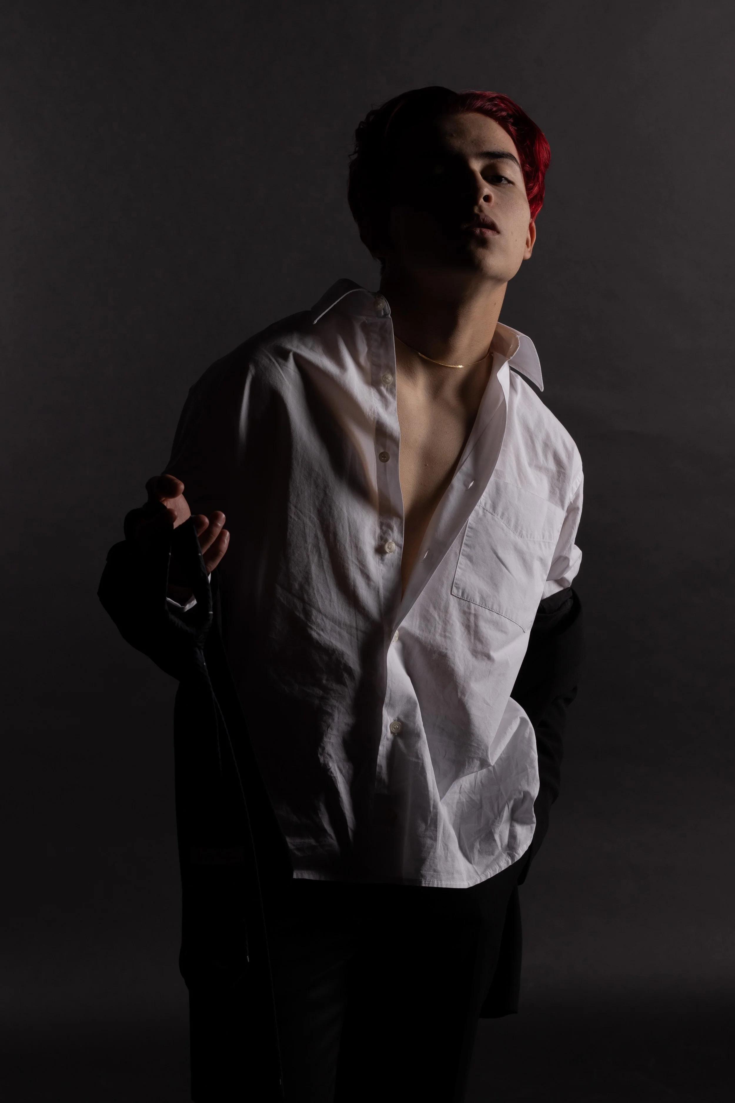 A person with red hair wearing a white shirt, partially unbuttoned, and black pants, standing against a dark background with low lighting.