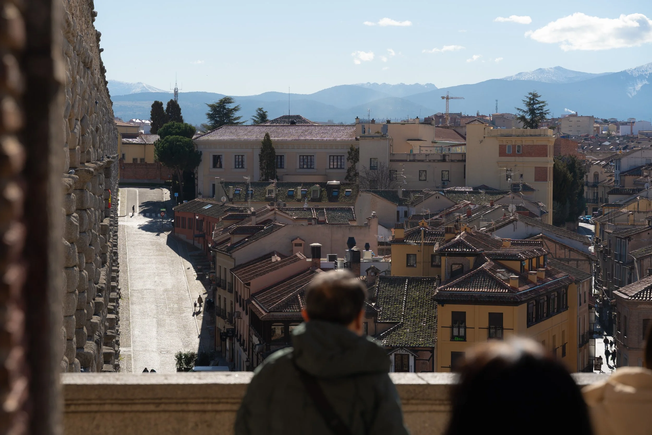 People sitting on a balcony overlooking a city with various buildings, rooftops, and distant mountains.