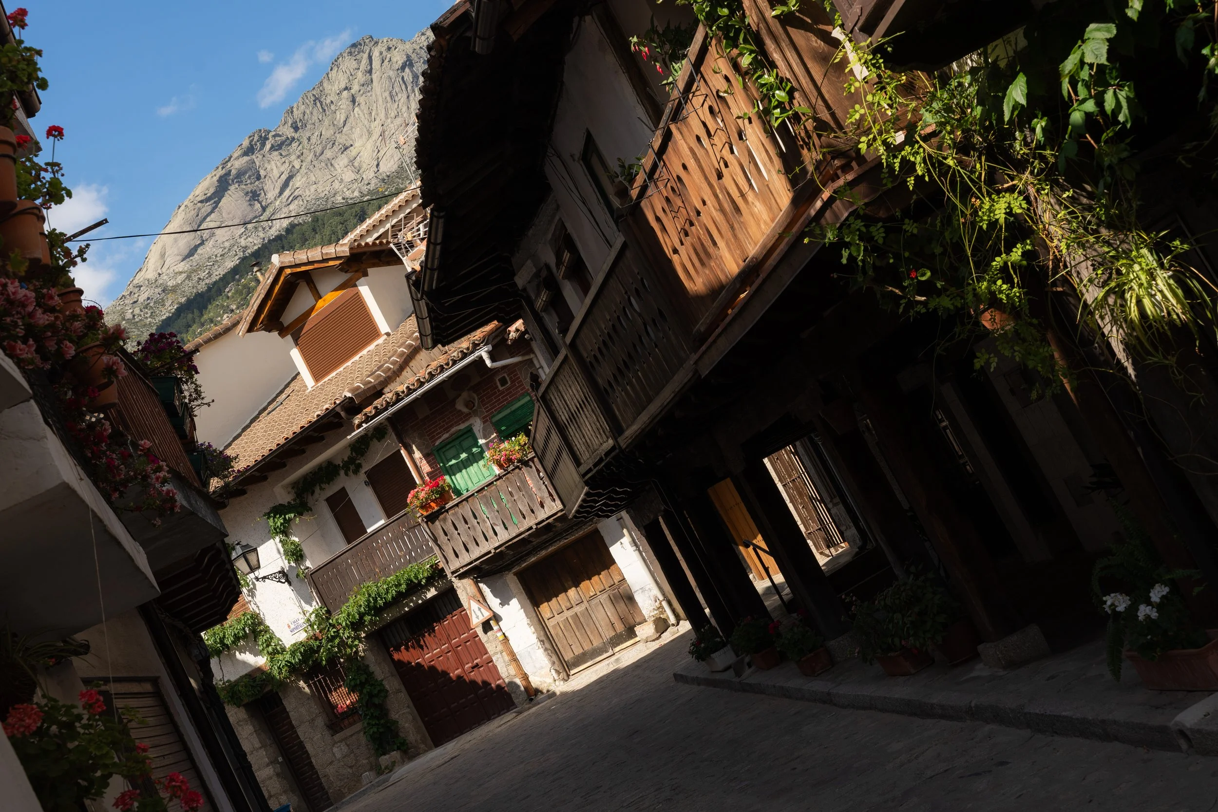 A narrow mountain village street lined with houses decorated with flower pots, wooden balconies, and shutters, with a rocky mountain in the background and a blue sky.