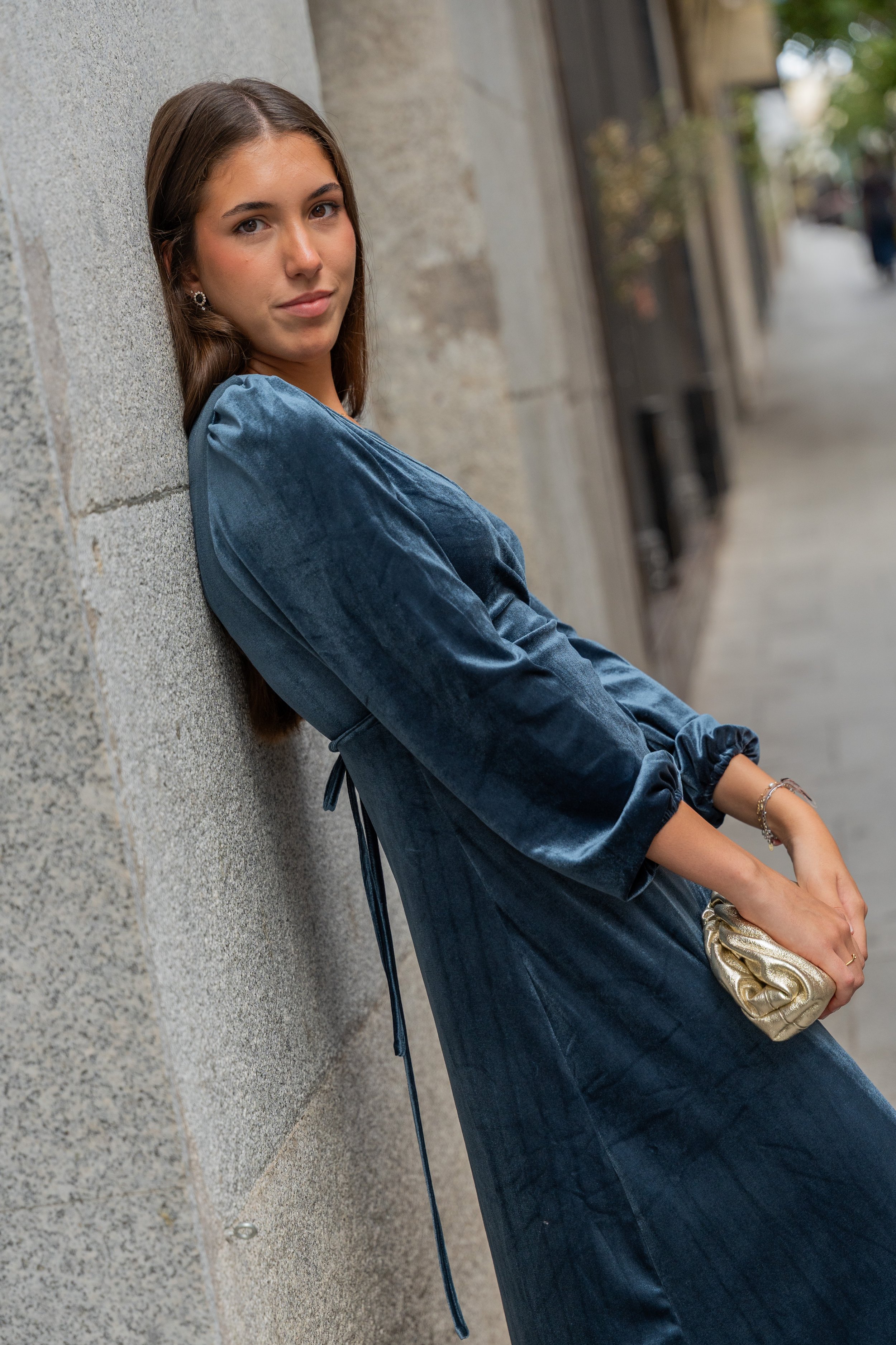 Young woman leaning against a stone wall on an urban sidewalk, wearing a blue velvet dress and holding a gold clutch.