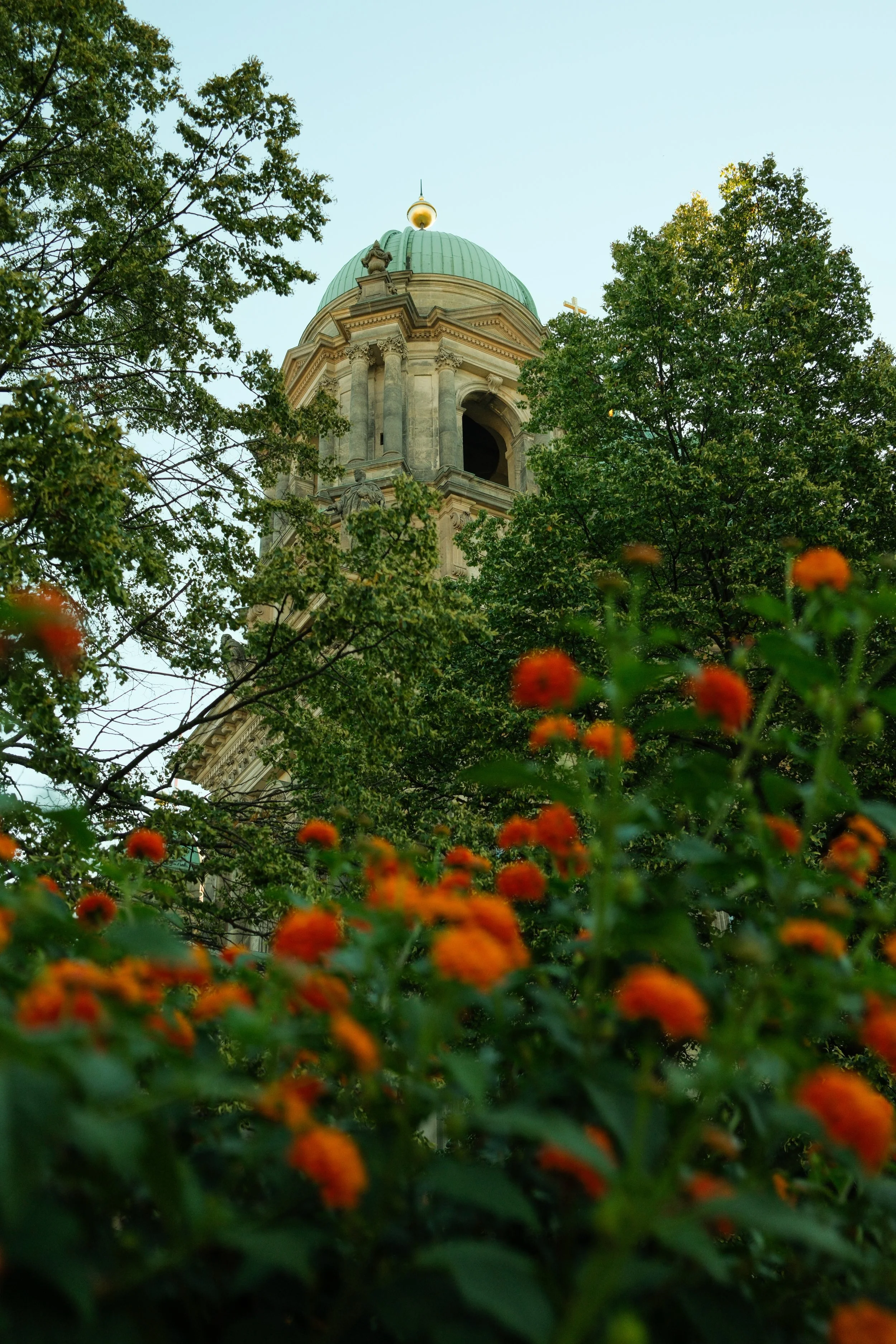 View of a historic building with a green dome and intricate architecture, partially obscured by green trees and orange flowers in the foreground.