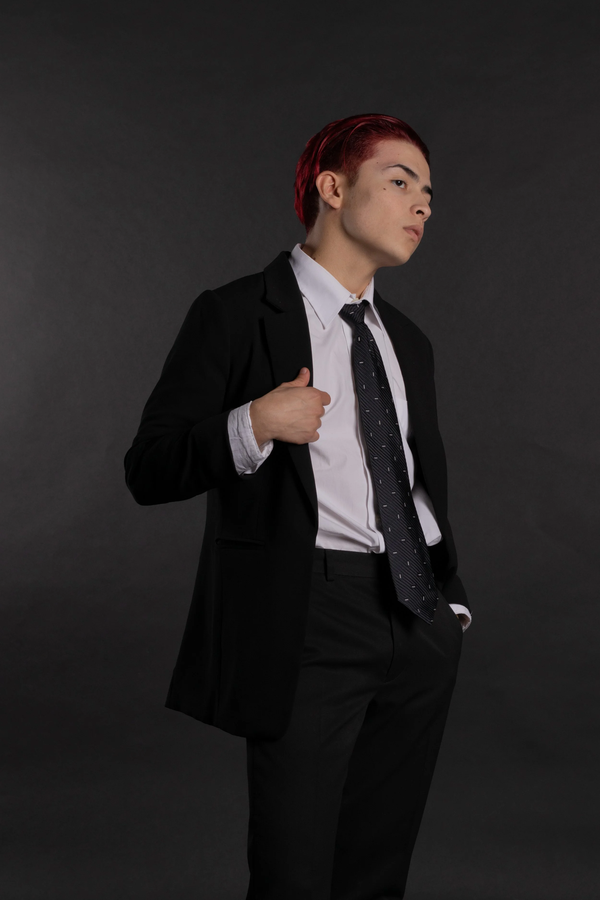 Young person with red hair wearing a black suit, white shirt, and black tie, posing against a dark background.