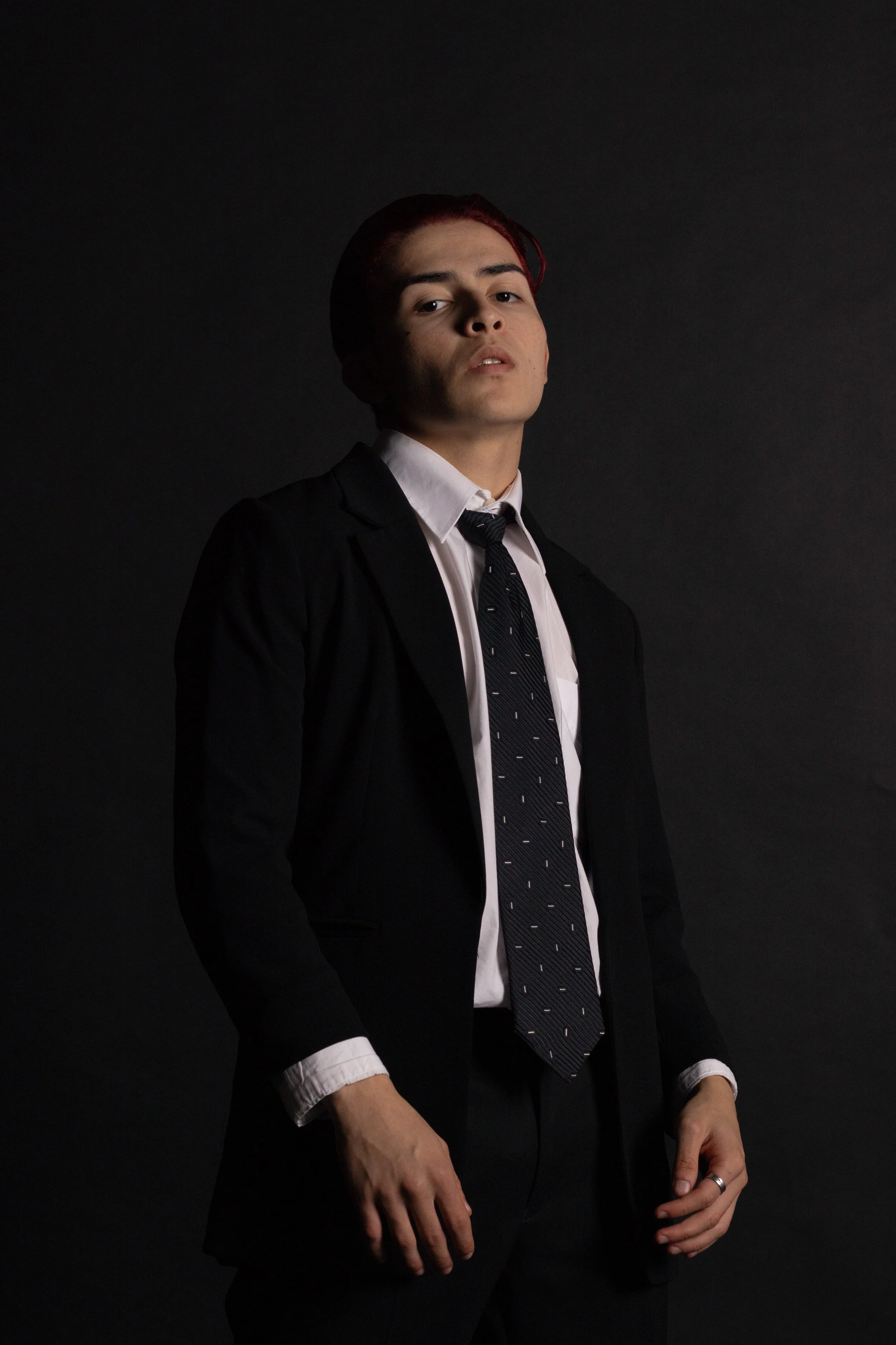 Young man with dyed red hair in a formal black suit, white shirt, and patterned tie, standing against a dark background with a serious expression.