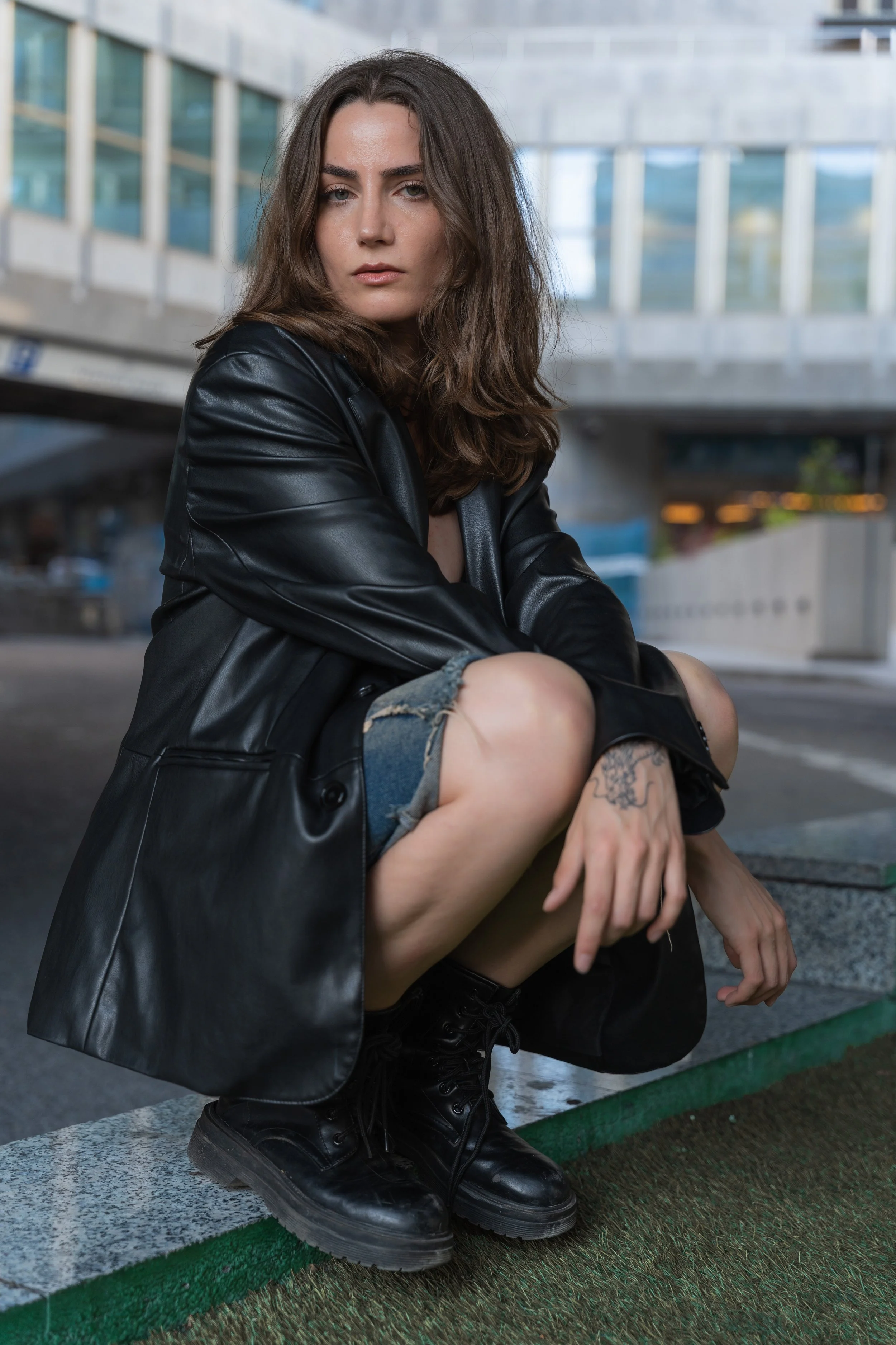 Young woman squatting outdoors in an urban setting, wearing a black leather jacket, ripped denim shorts, and black combat boots, with long brown hair and a tattoo on her hand.