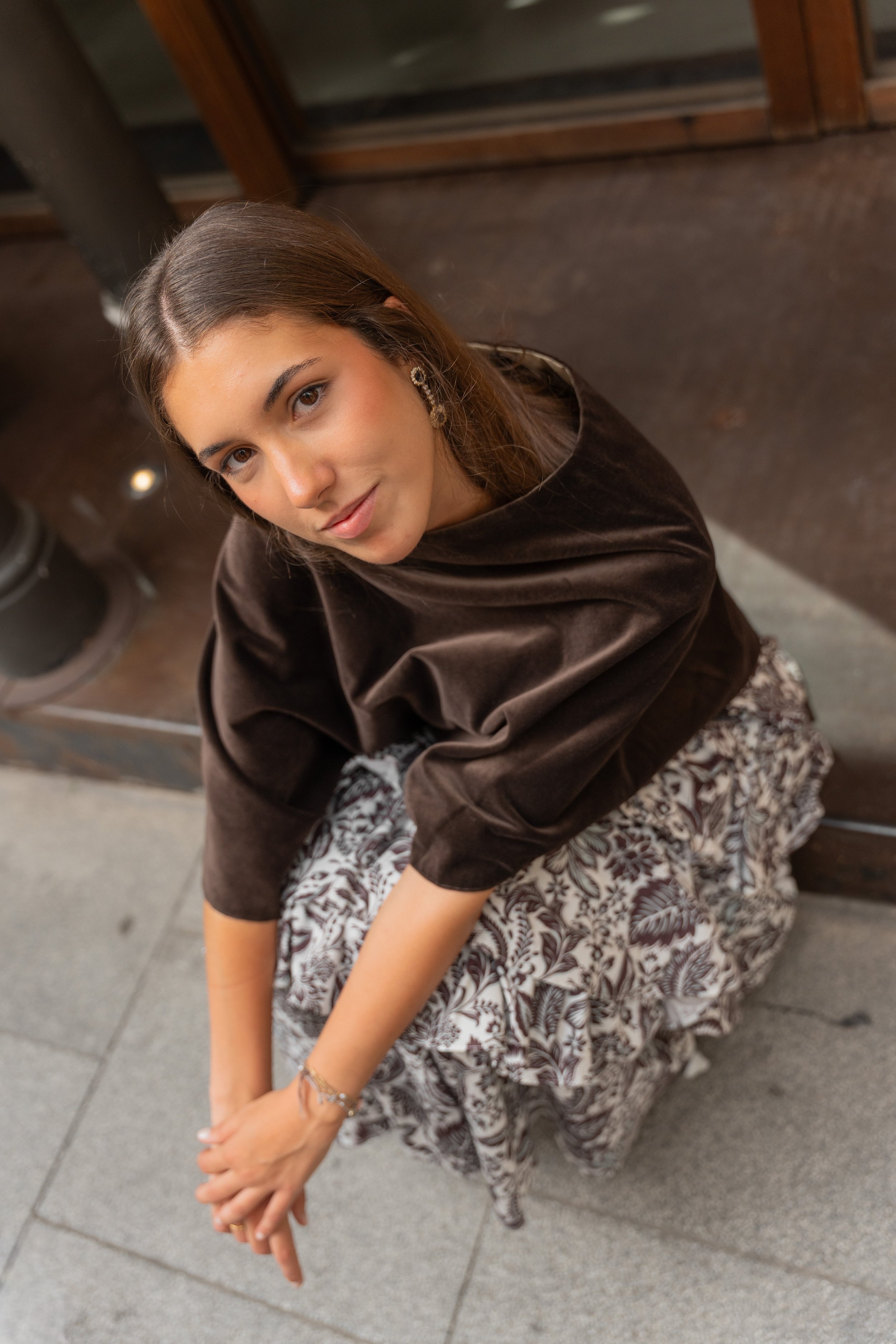 A young woman with long brown hair, wearing earrings, a black top with puffy sleeves, and a flowery skirt, is sitting on a bench, looking up with a slight smile.