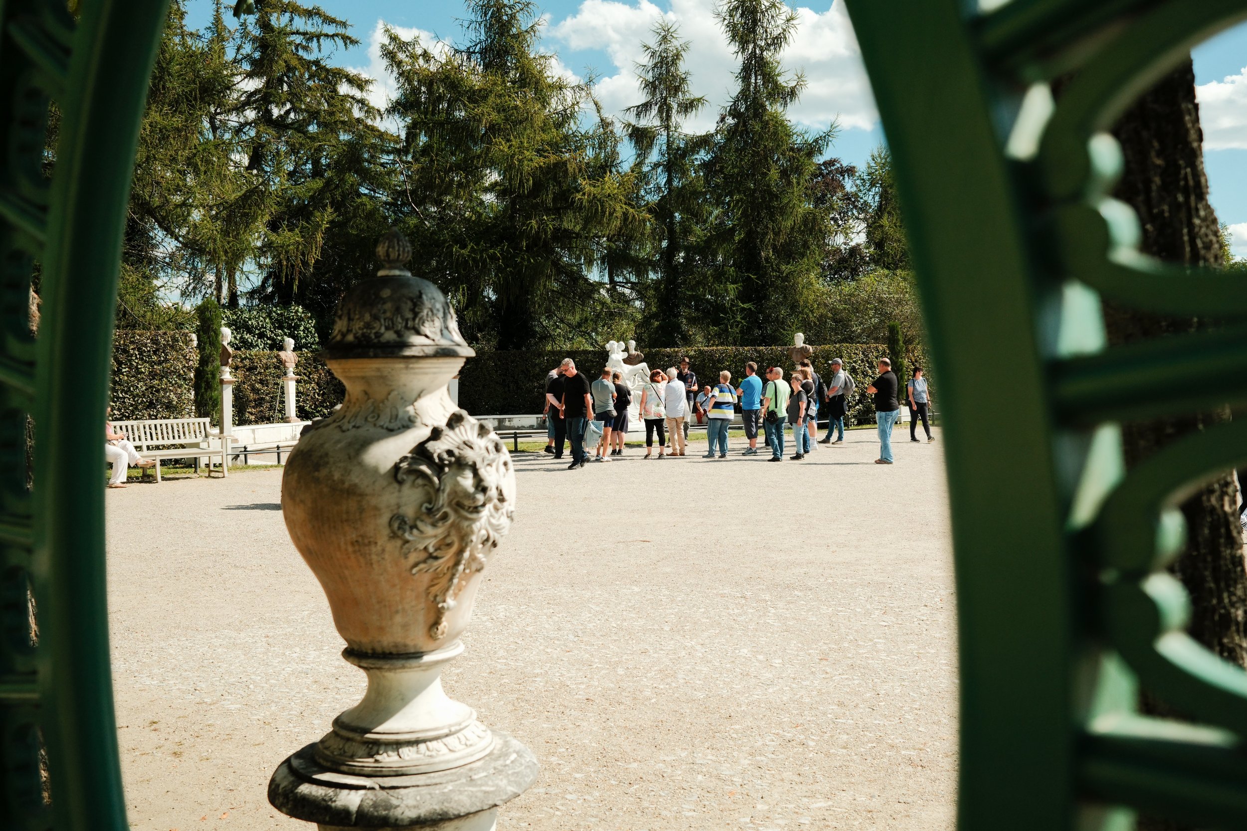 A group of tourists gathering in a courtyard, seen through green gate bars, with a decorative urn in the foreground and trees in the background.