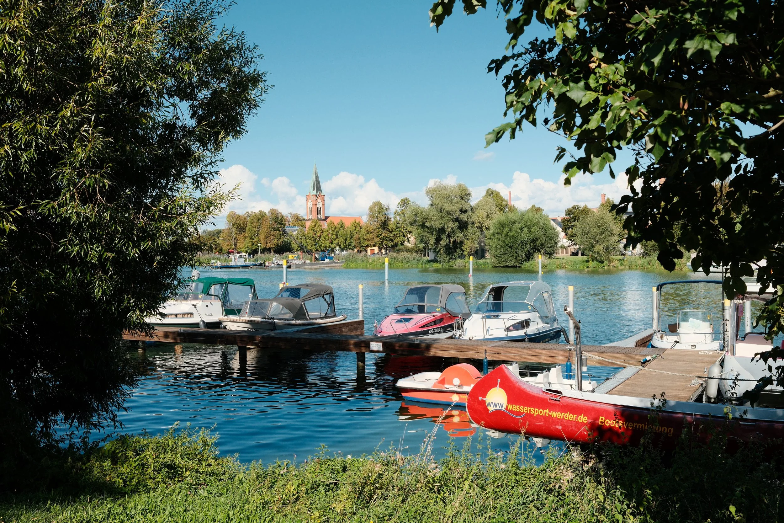 Boats docked at a marina on a calm river, with a town and church steeple in the background, surrounded by greenery on a sunny day.