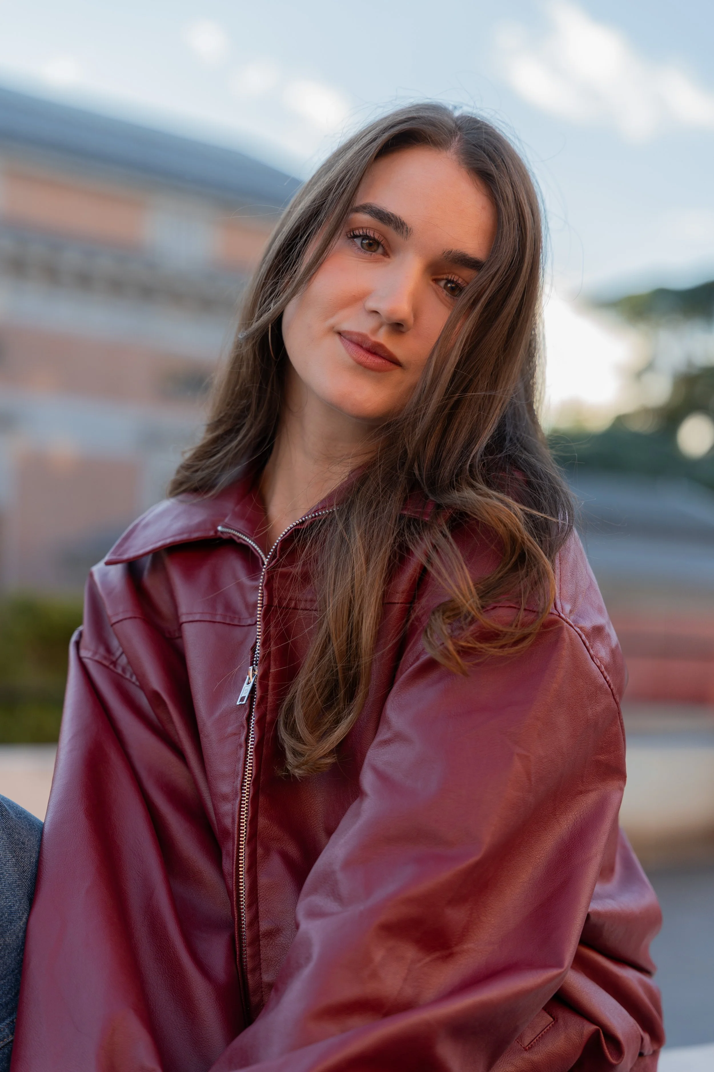 A young woman with long brown hair wearing a maroon leather jacket outdoors with a blurred background of buildings and trees.