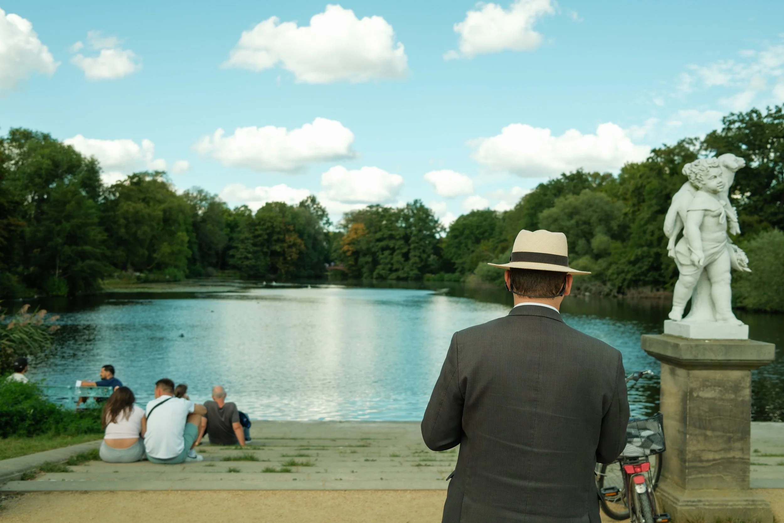 A man with a hat and suit stands with his back to the camera, looking at a lake with people sitting on a dock, surrounded by trees and a statue near the water.