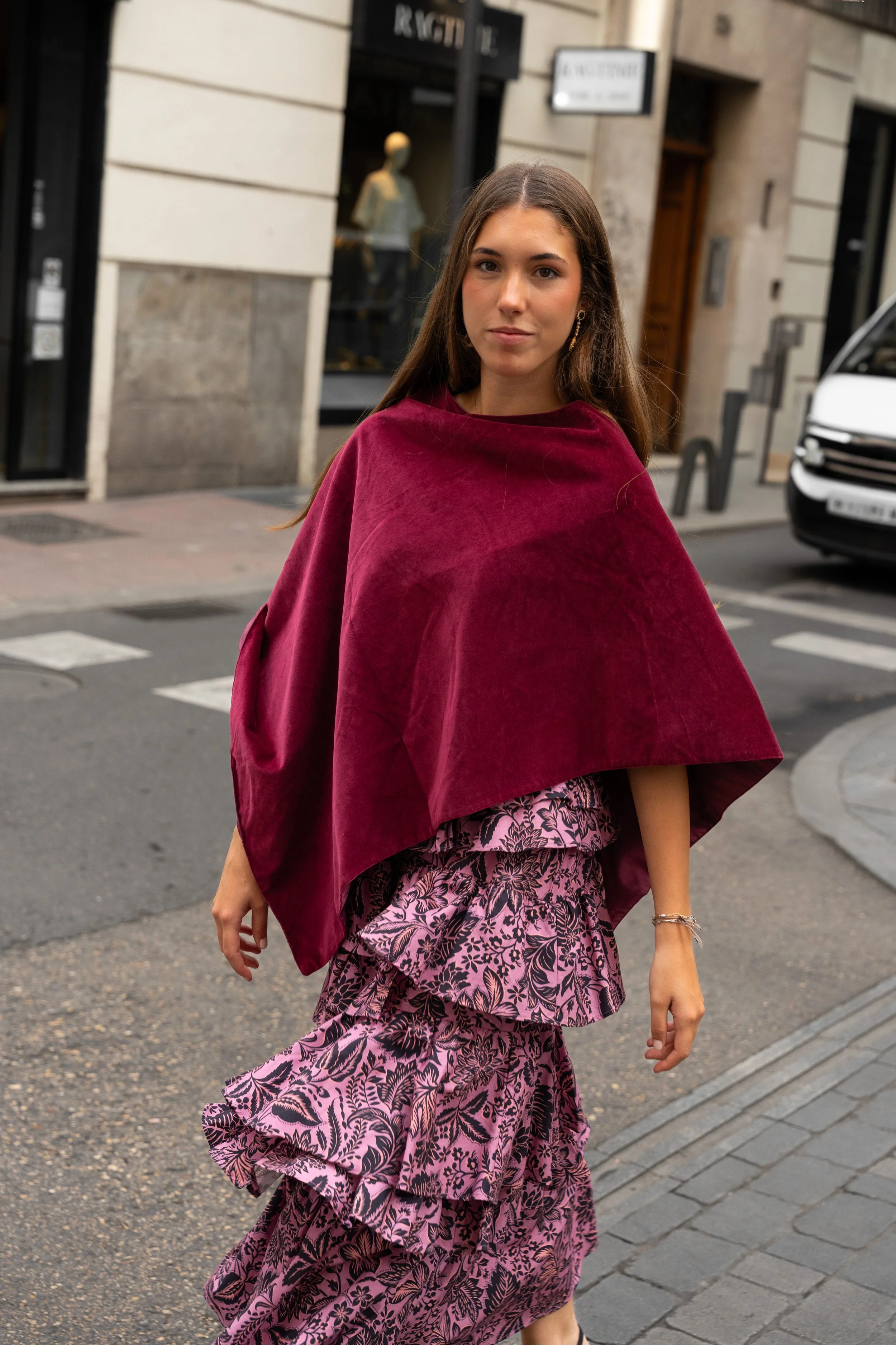 A young woman walking on a city street wearing a maroon cape and a pink and black floral layered dress.