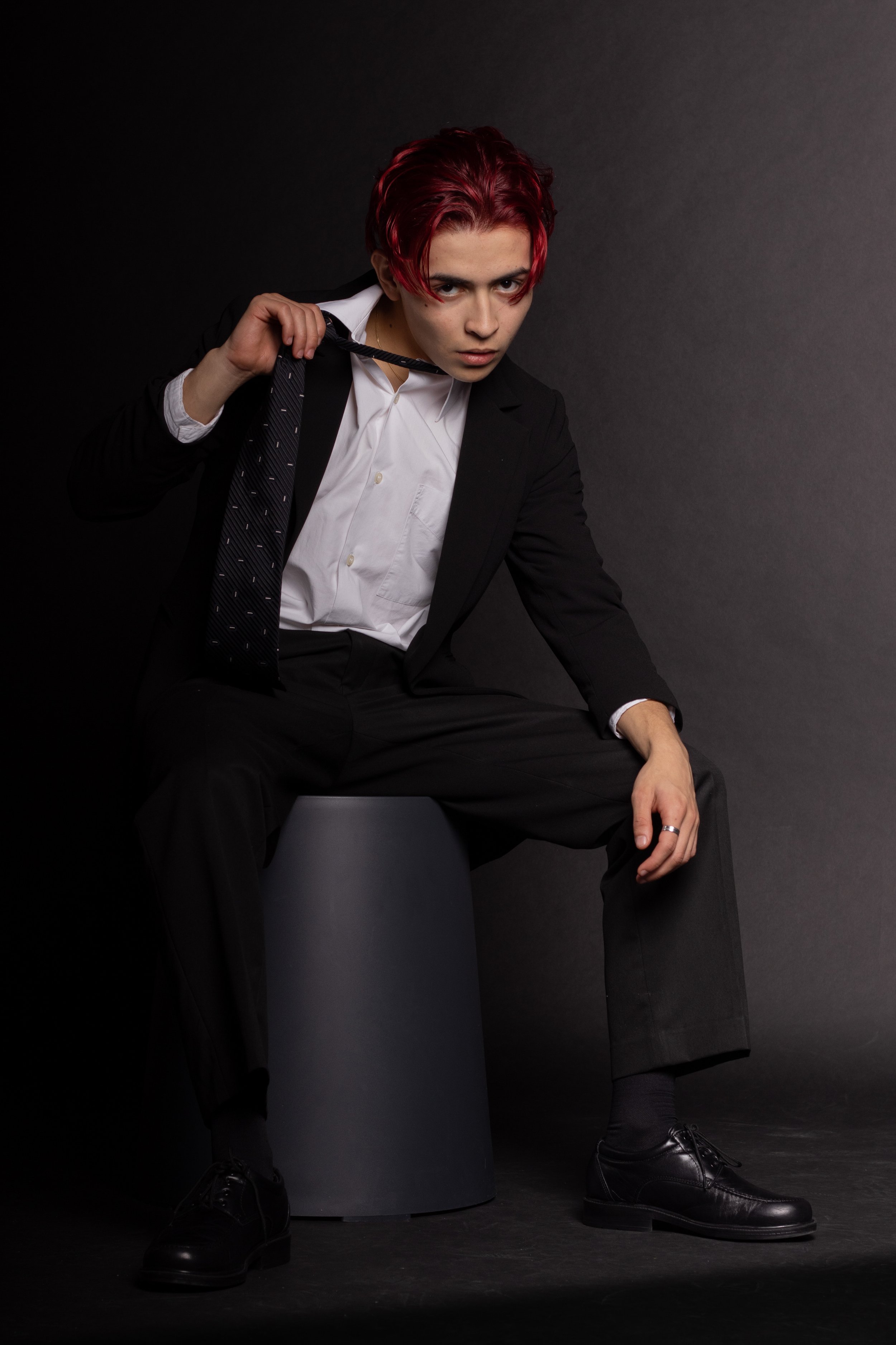Young man with red hair in formal black suit, white shirt, black tie, sitting on a gray stool against a black background, holding the tie with one hand.