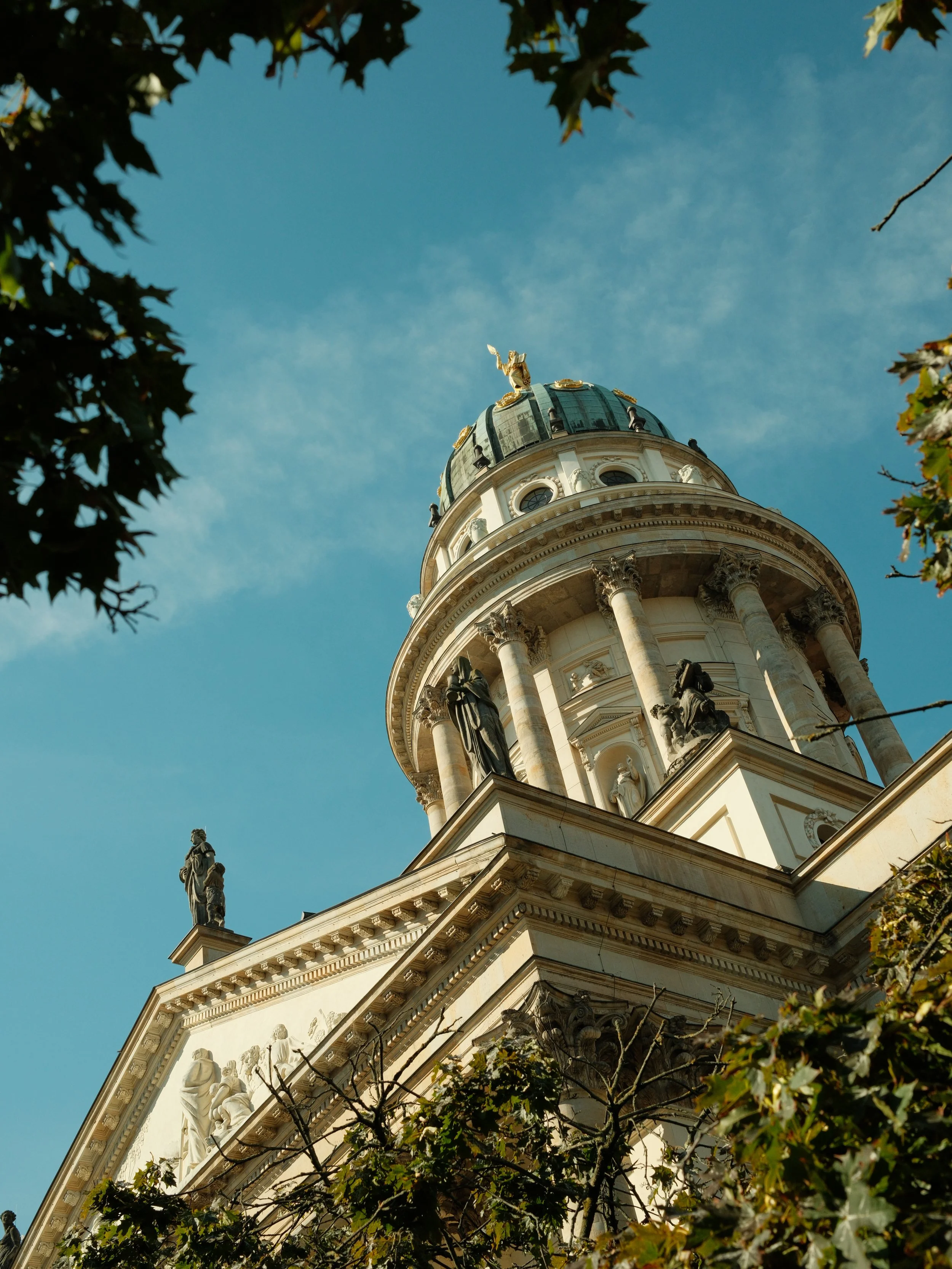 Looking up at a historic, ornate building with a gold statue on top, surrounded by trees with green leaves, under a blue sky.