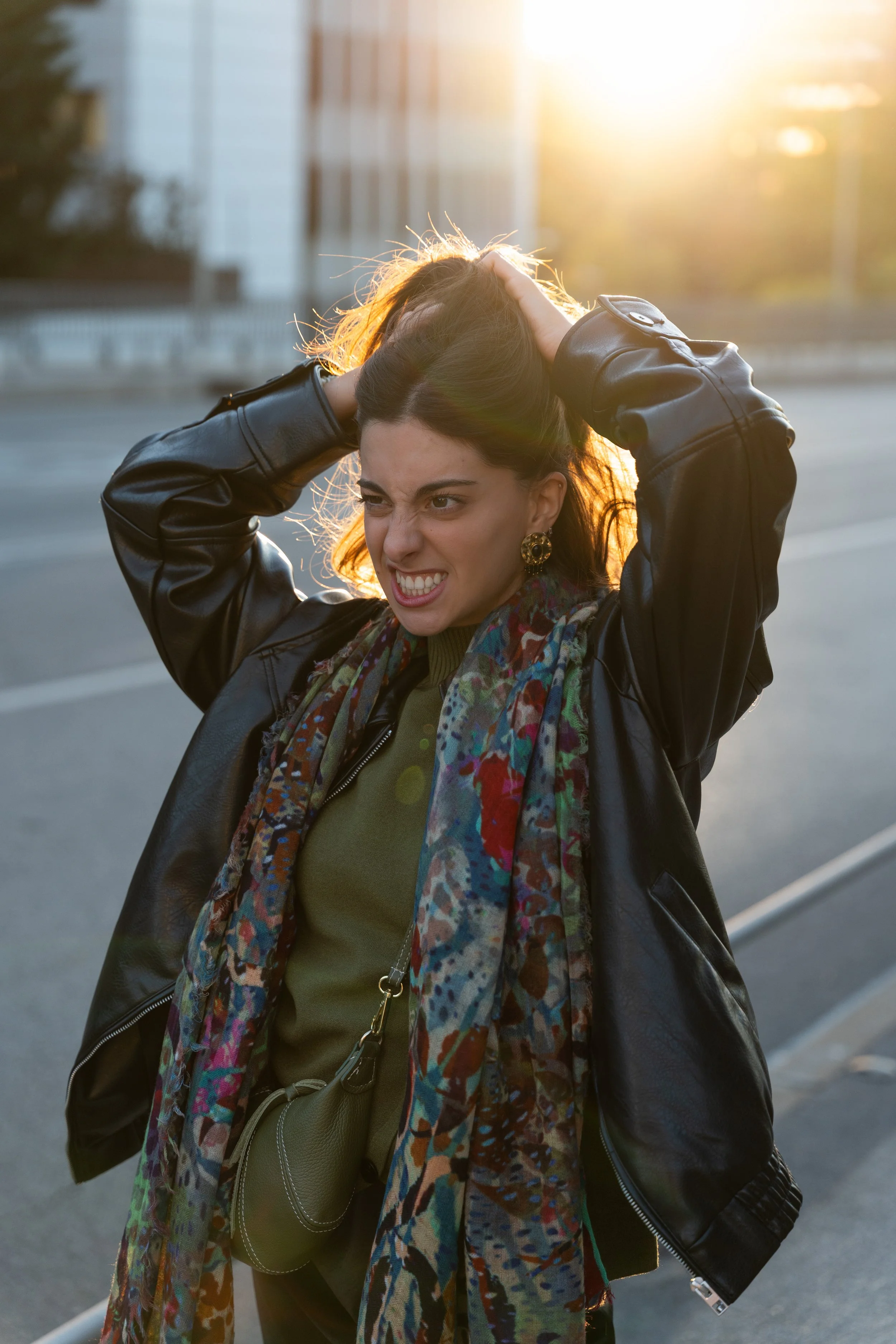 Young woman wearing a black leather jacket and colorful scarf, pulling her hair back, with a sun flare in the background on an urban street.
