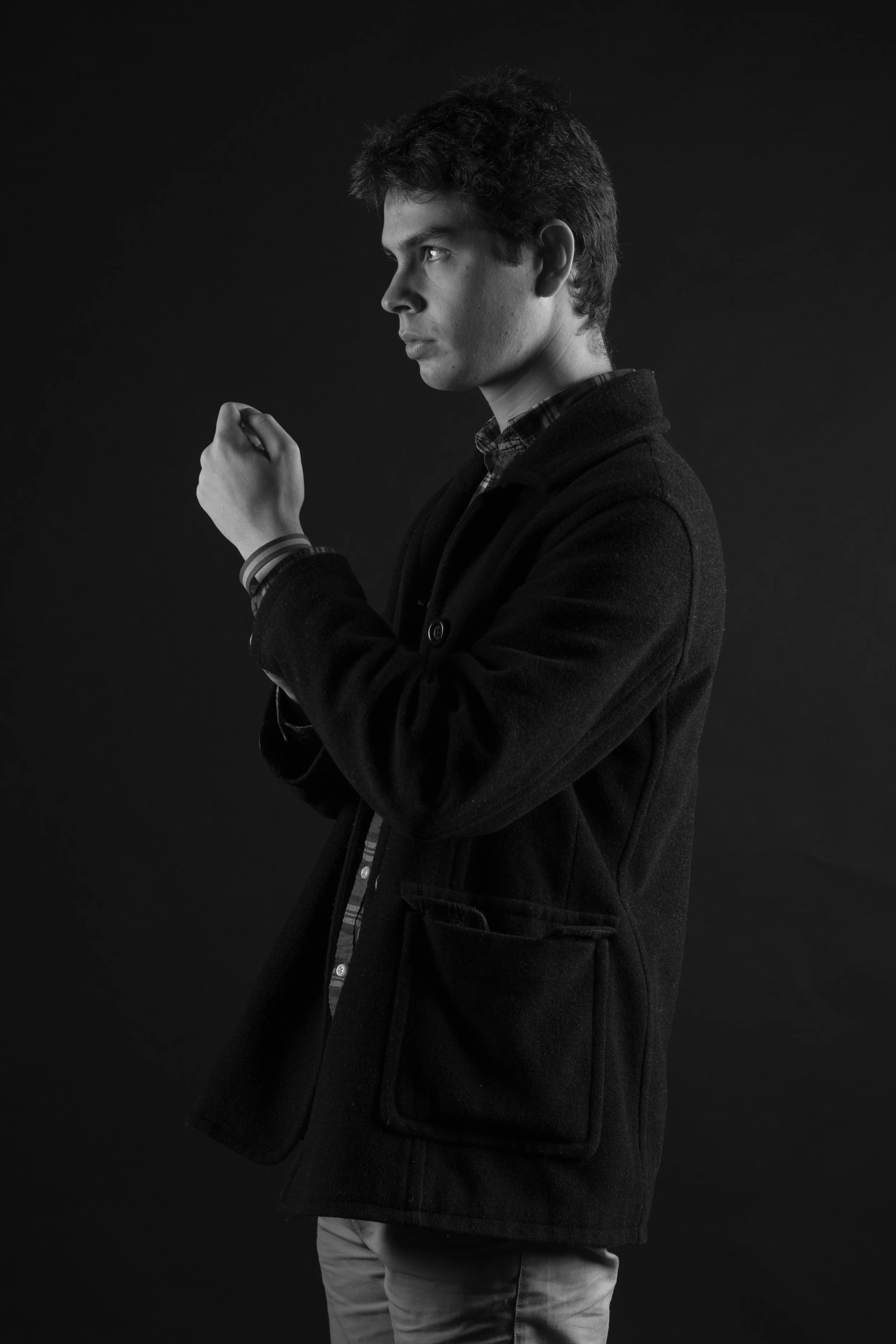 Black-and-white portrait of a young man with dark, tousled hair, dressed in a dark coat, looking to the side, against a dark background.