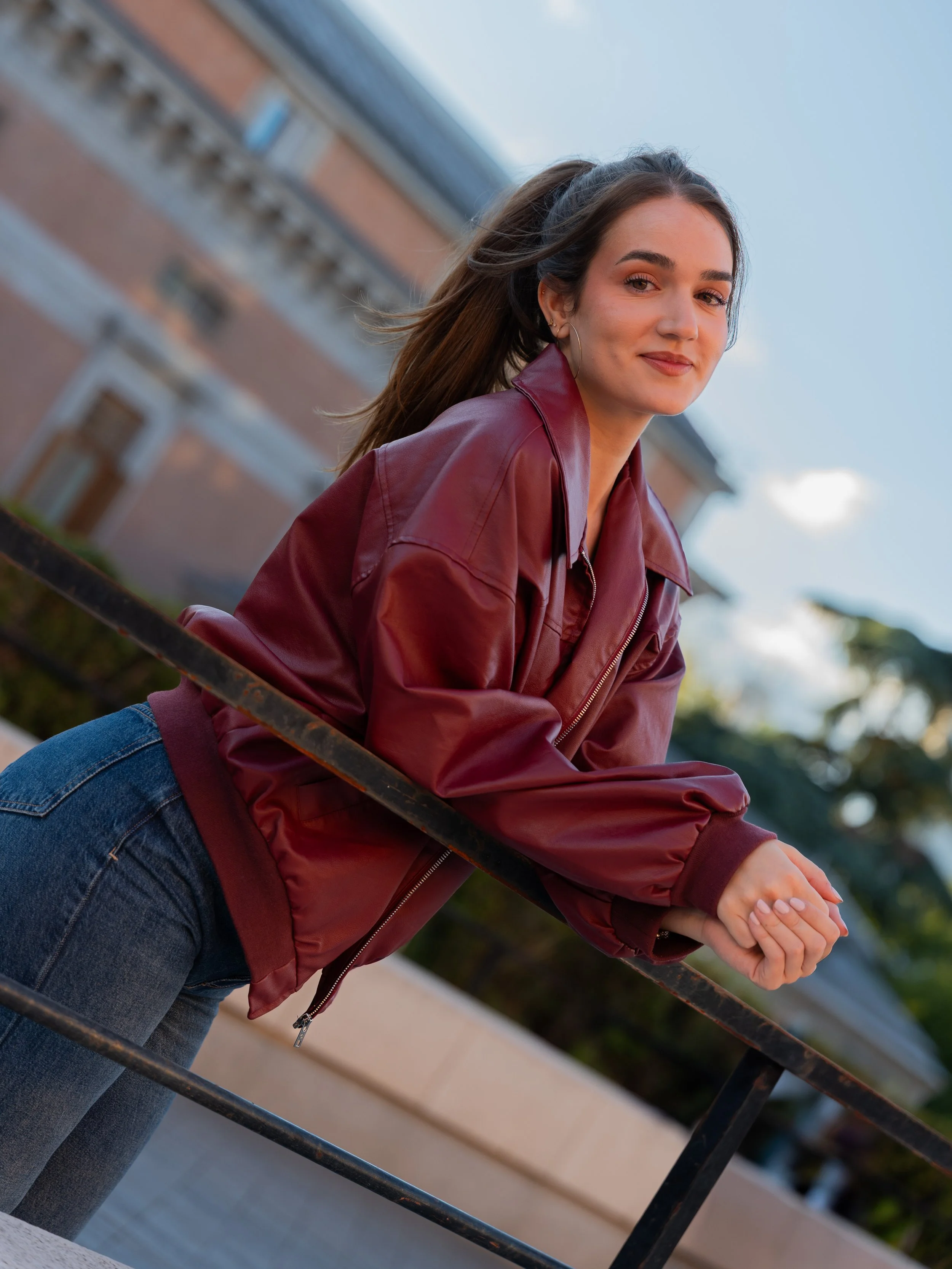 Young woman with long brown hair wearing a burgundy leather jacket and jeans, leaning on a black metal railing outdoors with a brick building and trees in the background.