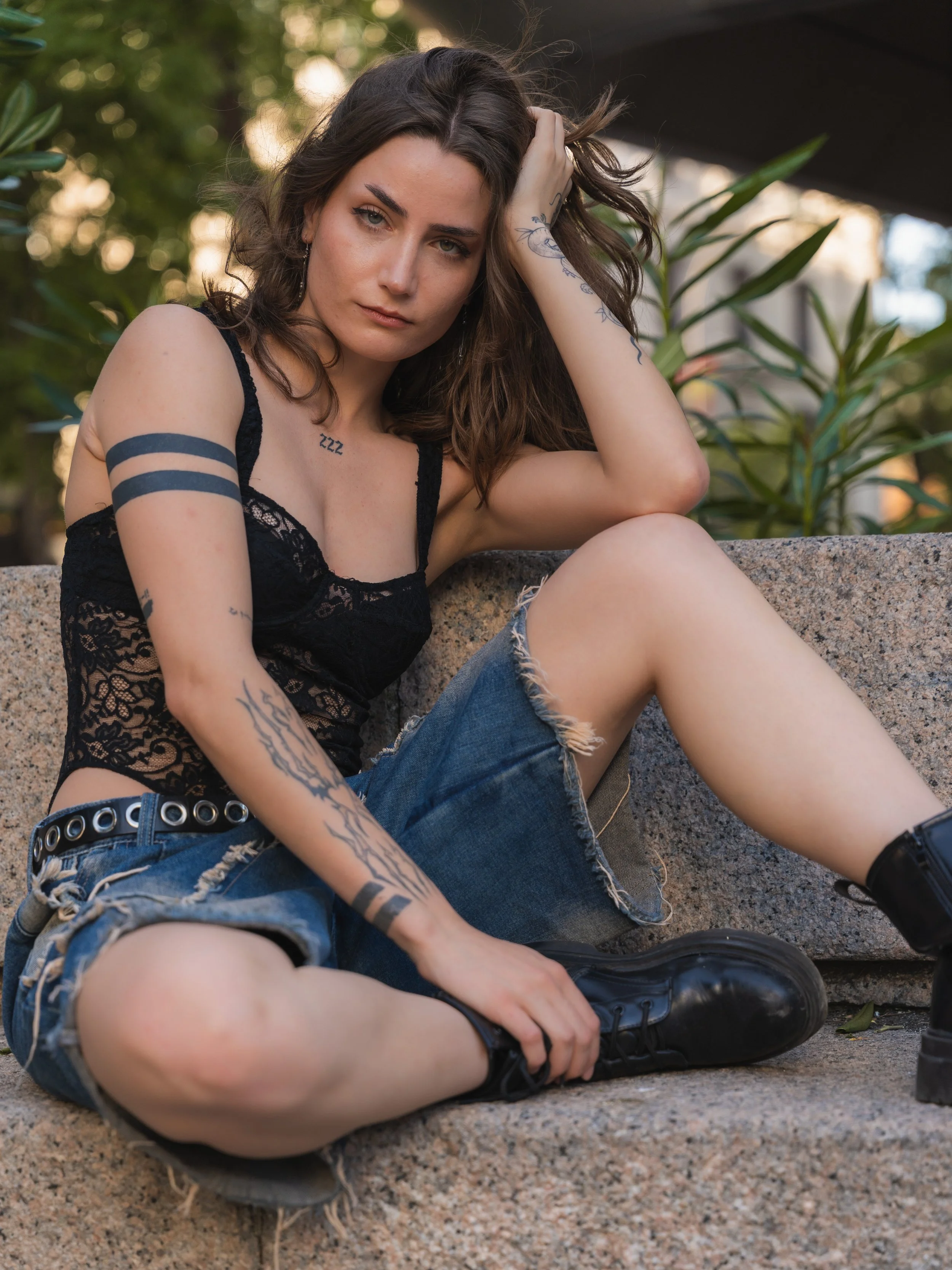 A young woman with tattoos, wearing a black lace crop top, ripped denim shorts, and black boots, sitting on a stone bench outdoors, looking at the camera.