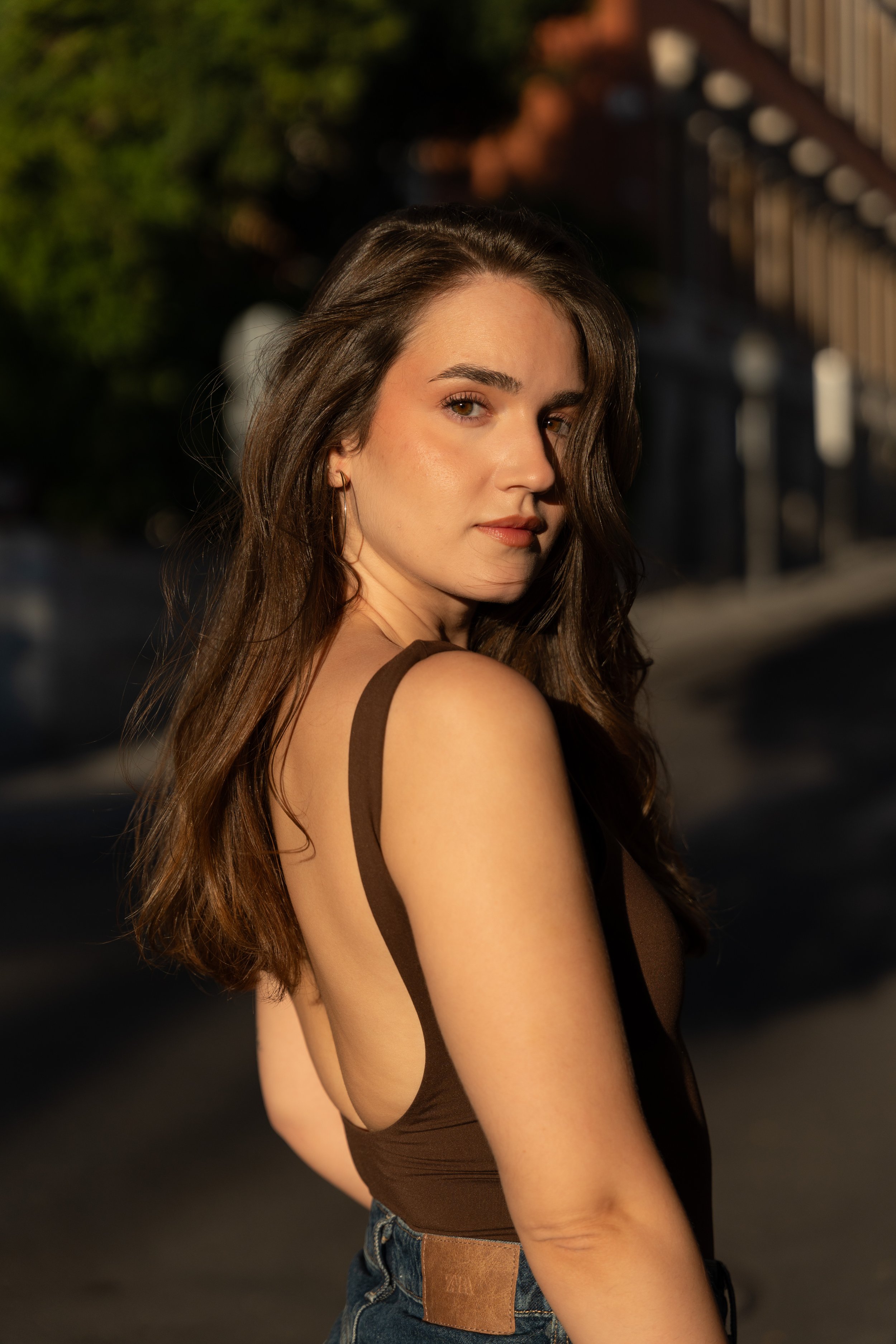 Young woman with long wavy brown hair, wearing a brown sleeveless top and blue jeans, standing outside during golden hour with sunlight illuminating her face and shoulder.