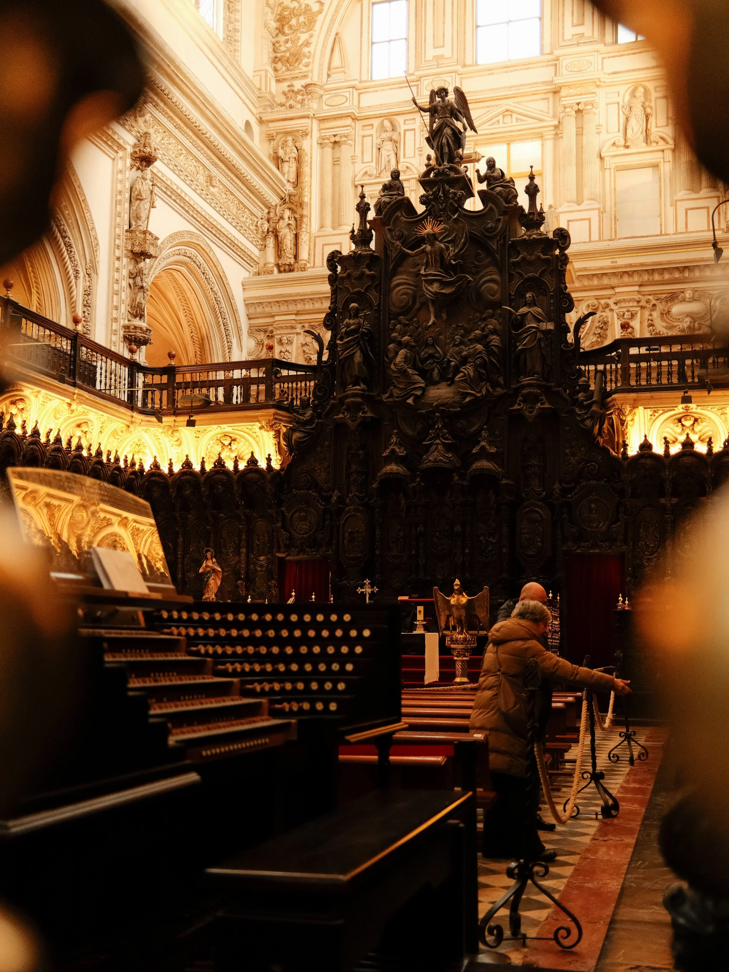 Inside a historic church, a person in a brown coat is praying near an altar, with a large pipe organ in the foreground and ornate woodwork and statues in the background.