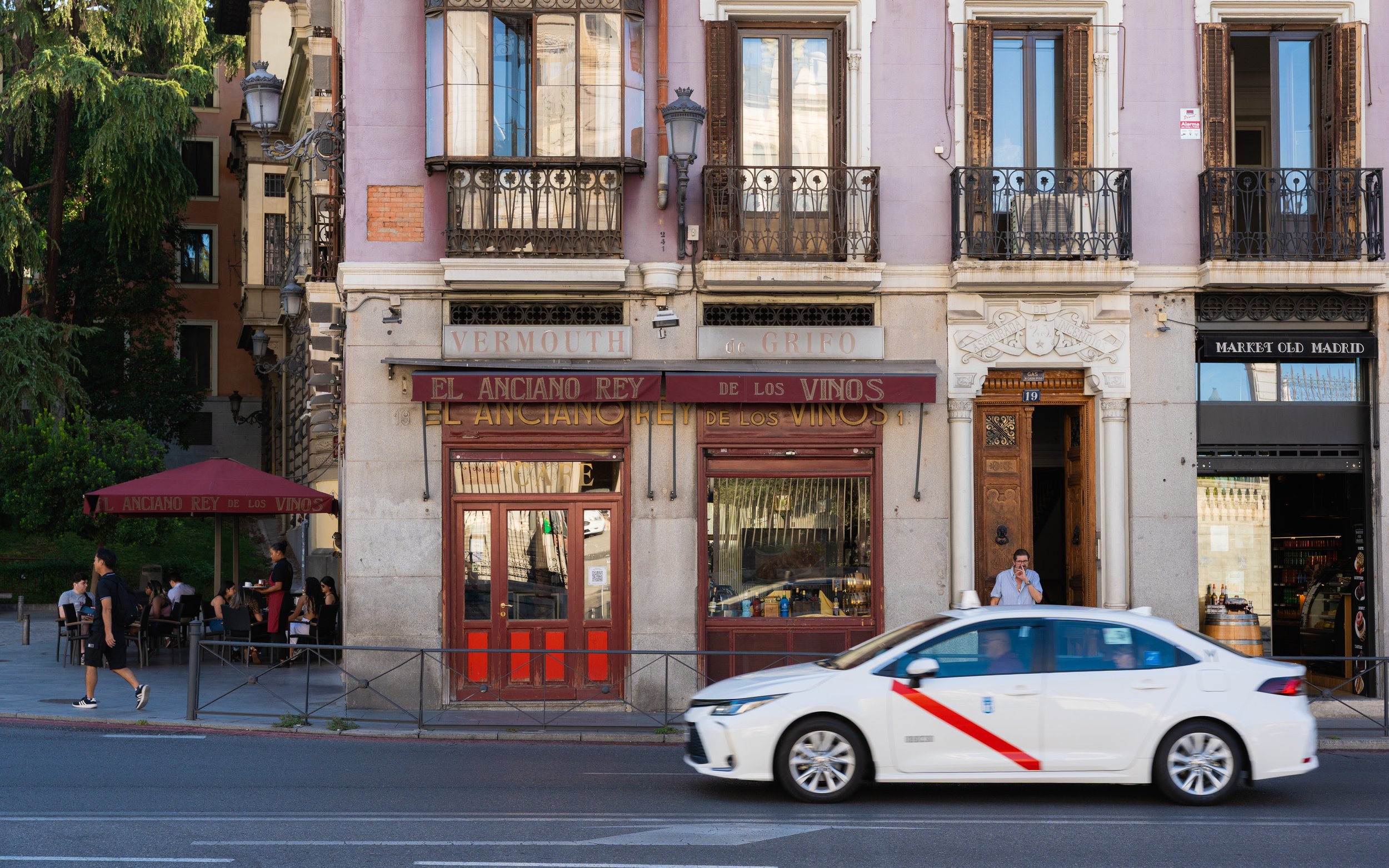 Street view of a building with a restaurant on the ground floor and people sitting outside under a red umbrella. A white car is driving by in the foreground.