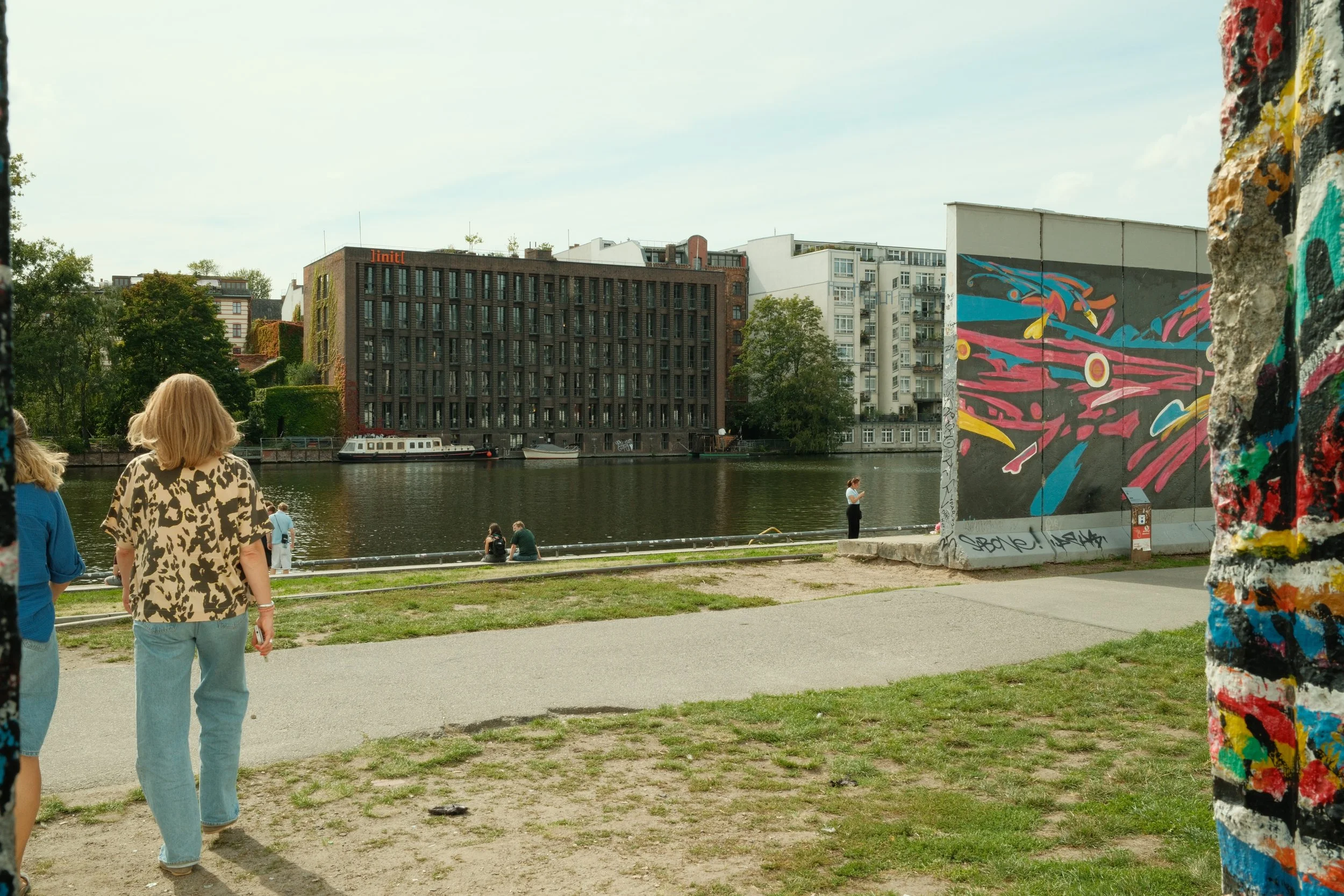 People walking along a riverside path near a bridge with graffiti-covered walls, with buildings and trees in the background.