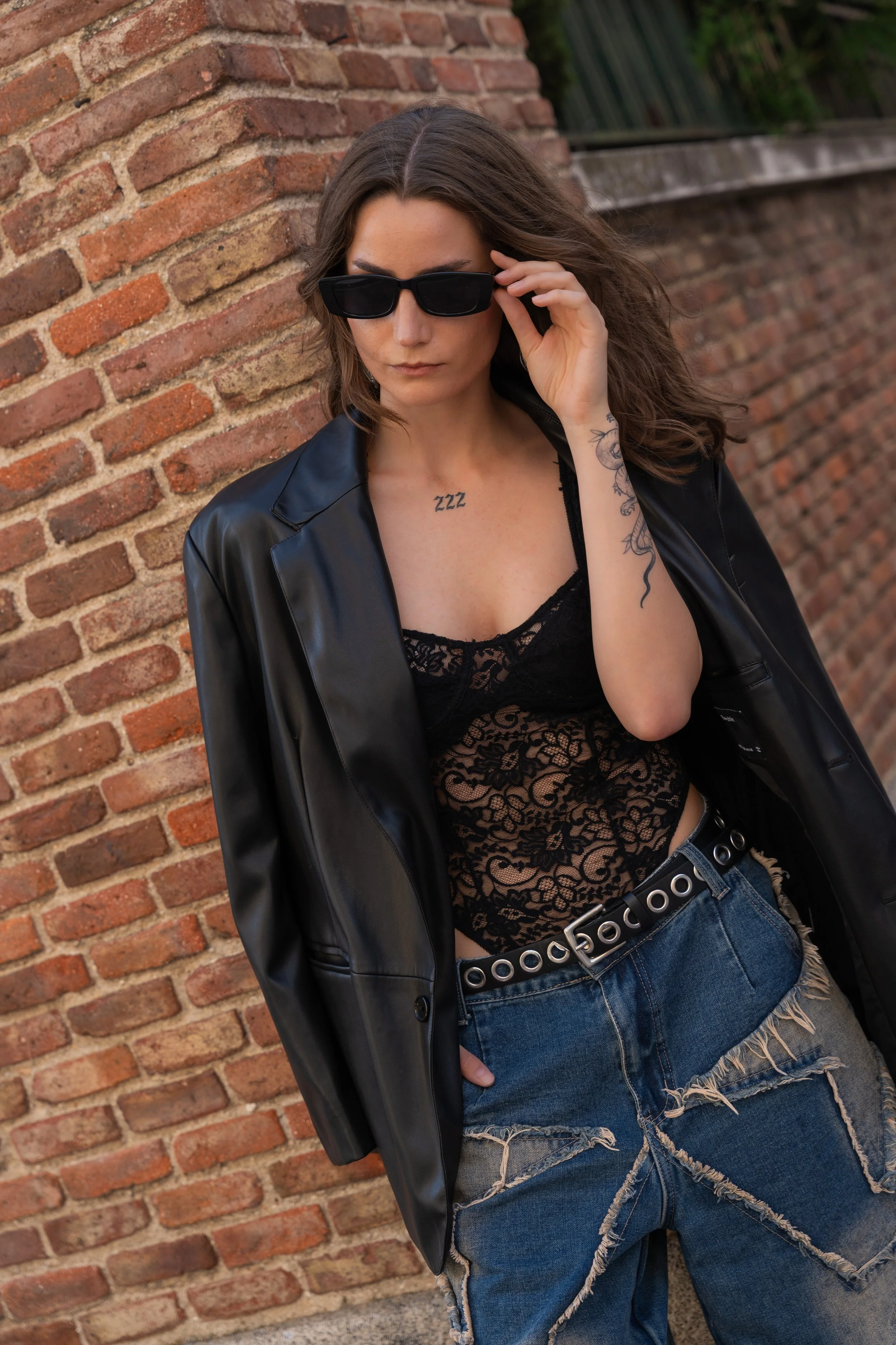 A woman with long brown hair wearing black sunglasses, a black lace top, a black leather jacket draped over her shoulders, and baggy distressed jeans with a wide belt, standing against a brick wall, looking slightly downward and touching her sunglass