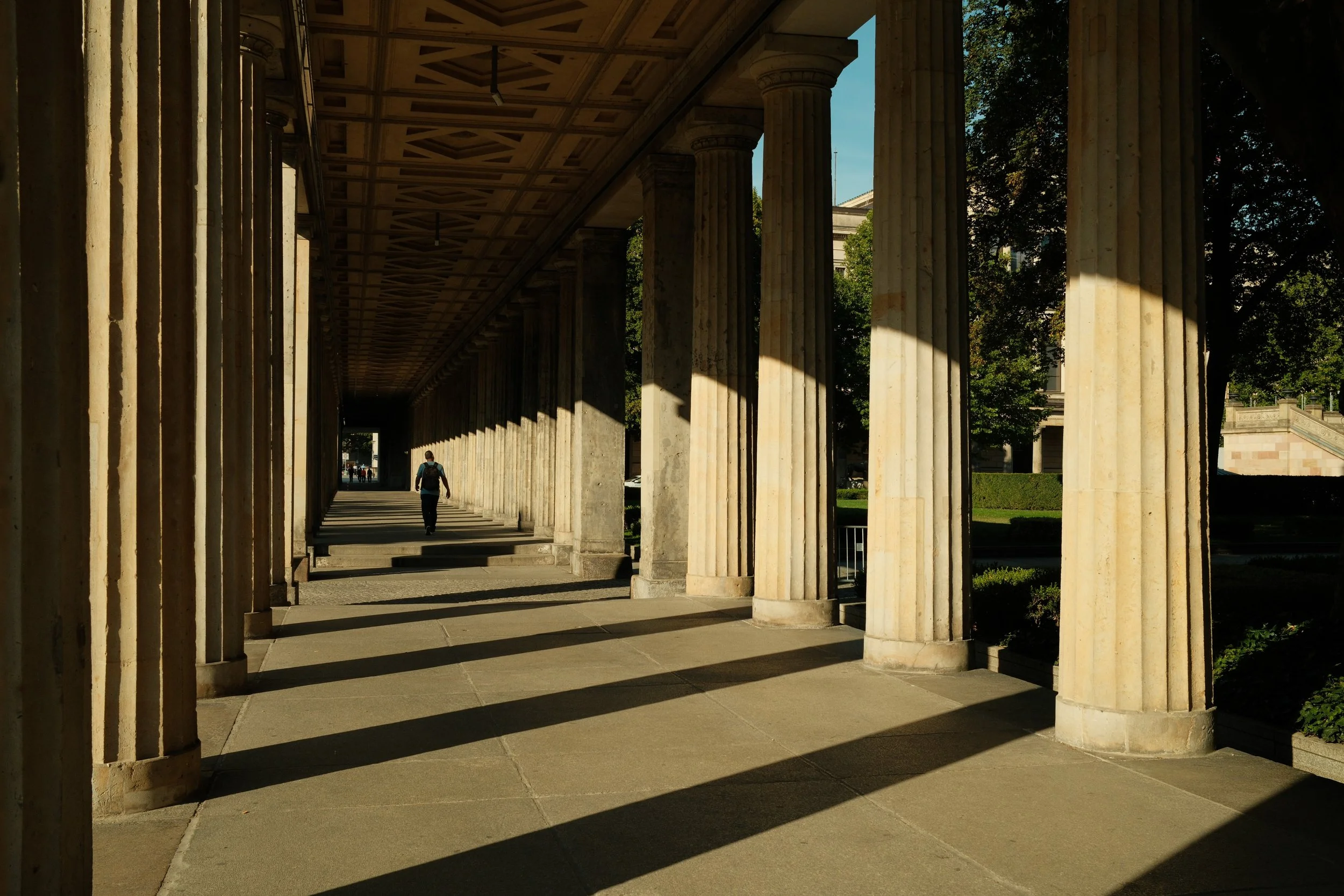 A person walking along a colonnade with classical columns, casting long shadows on the ground, in a park or historic site during late afternoon.