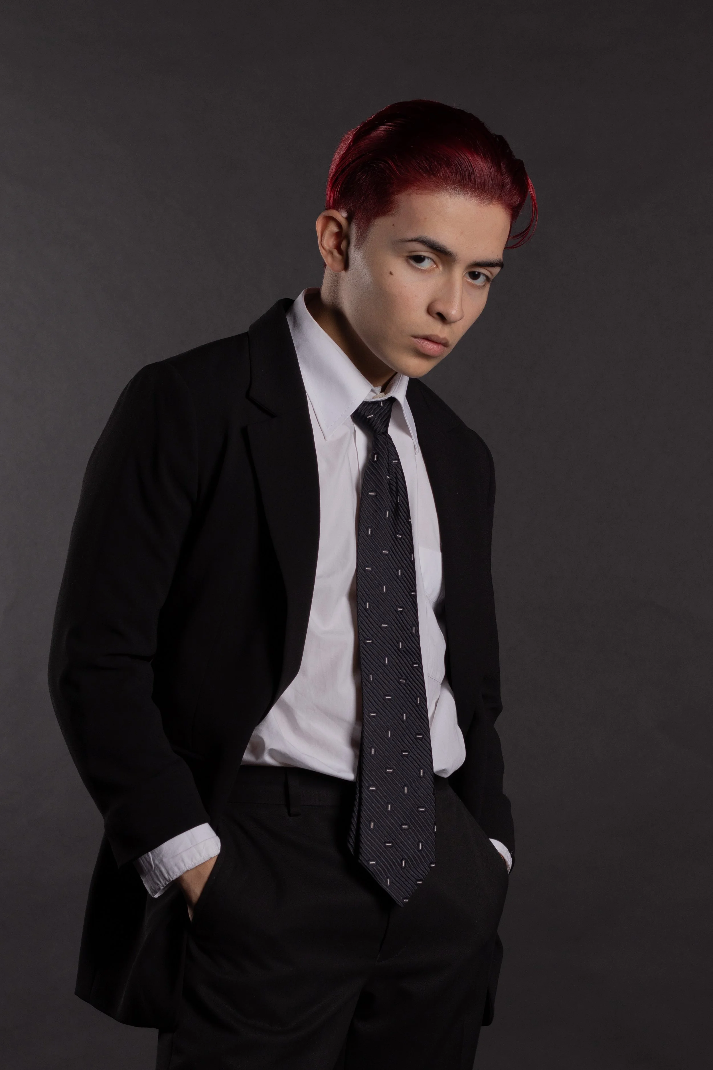 Young person with short red hair wearing a black suit, white shirt, and black tie with small white patterns, posing against a dark gray background.