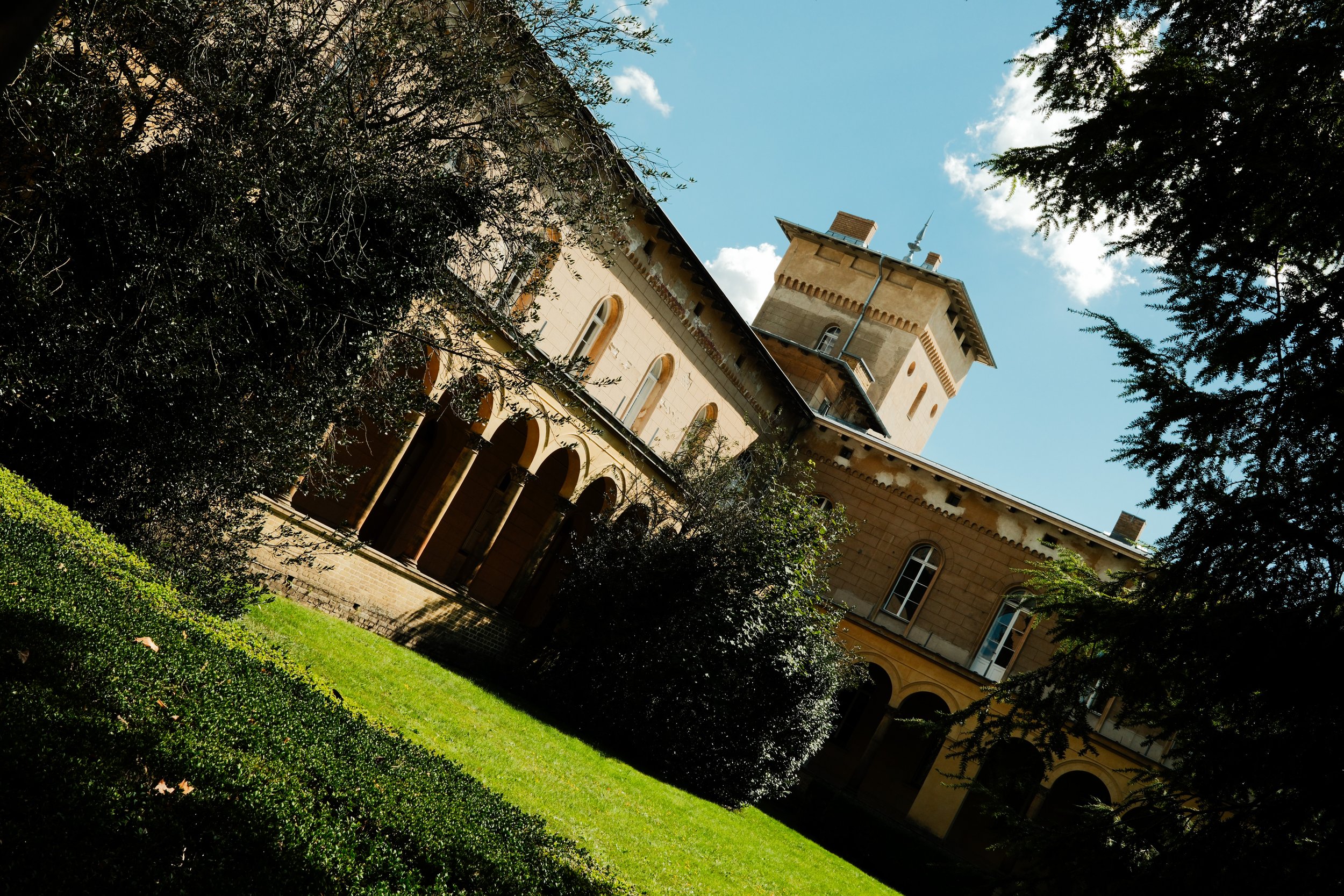 A historic beige castle-like building with arched windows, surrounded by green trees and grass, under a blue sky with some white clouds.