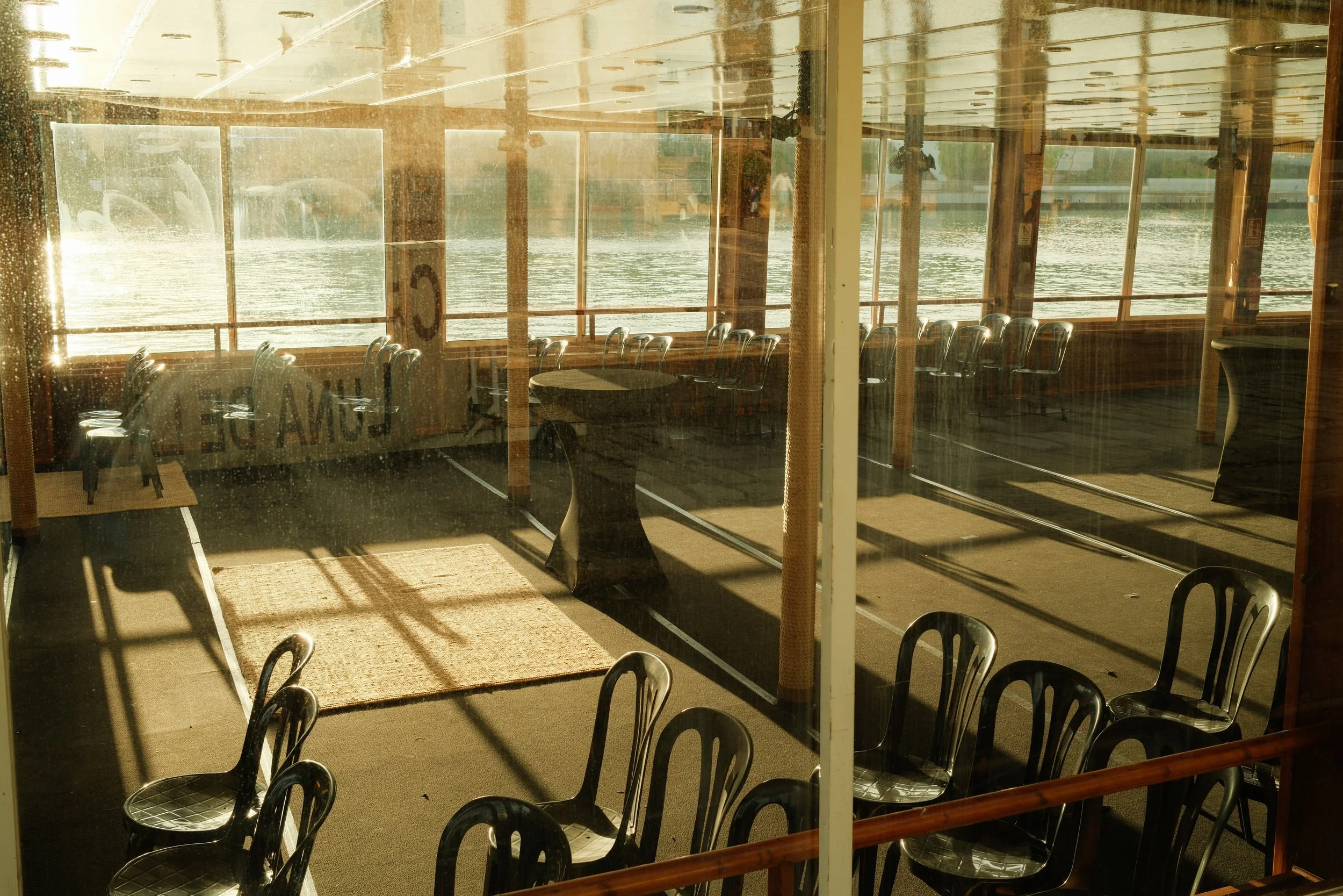 Empty indoor seating area with black chairs, tables, and large windows overlooking a river at sunset.